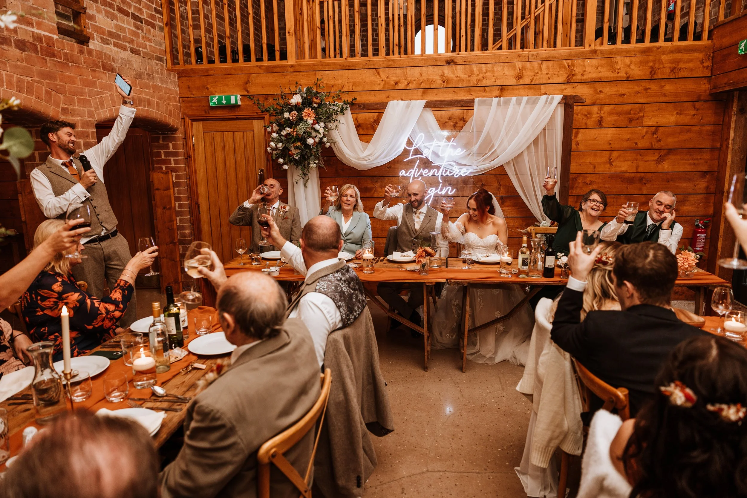 A wedding celebration in a rustic indoor venue with wooden walls and brick accents. The bride and groom sit at the head table raising glasses, surrounded by friends and family. A man standing on the left is giving a toast with a microphone. There is 