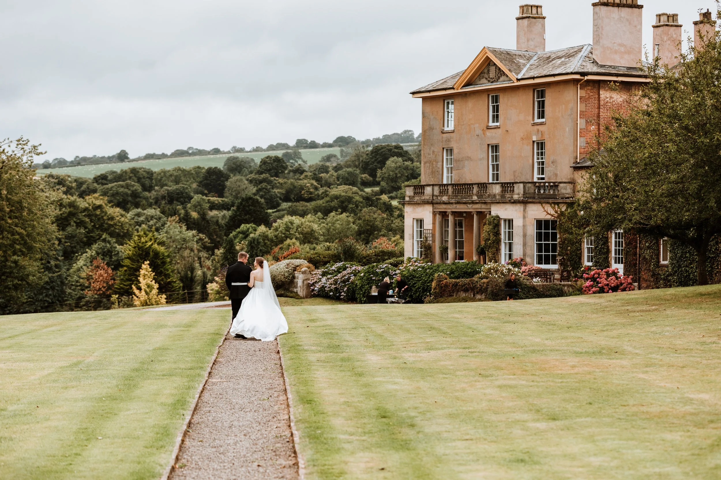 A bride and groom holding hands walking on a gravel path in front of a large, historic house surrounded by a well-maintained garden with colorful flowers and greenery.
