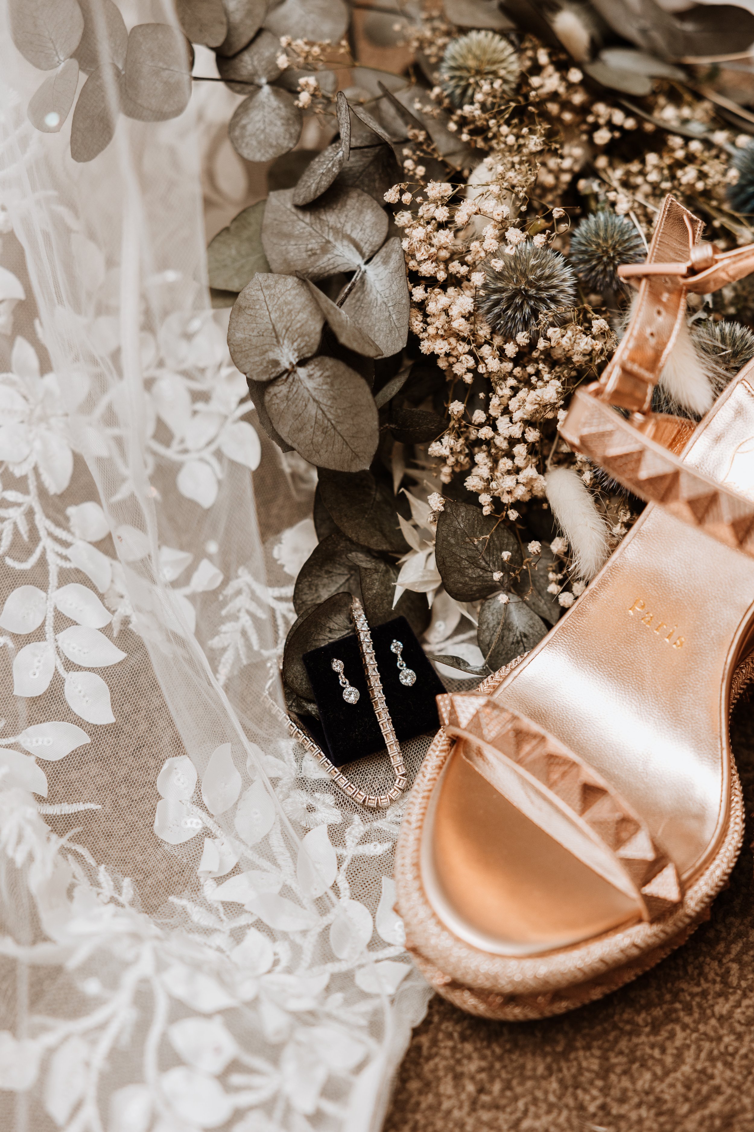 A collection of jewelry, including earrings and a necklace, displayed in front of a floral arrangement with eucalyptus leaves and small flowers, alongside a pair of metallic pink shoes.