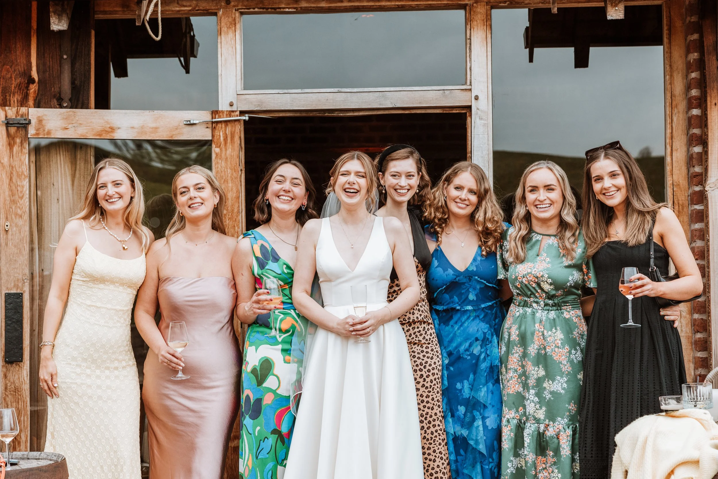 Group of women dressed in formal attire at a celebration, standing in front of a wooden structure, some holding glasses of wine, smiling for the camera.