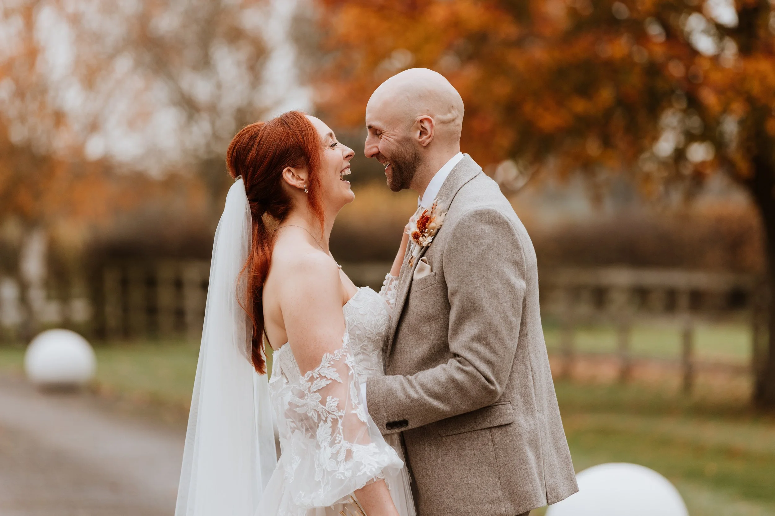 A bride and groom smiling and looking at each other outdoors with autumn trees in the background.