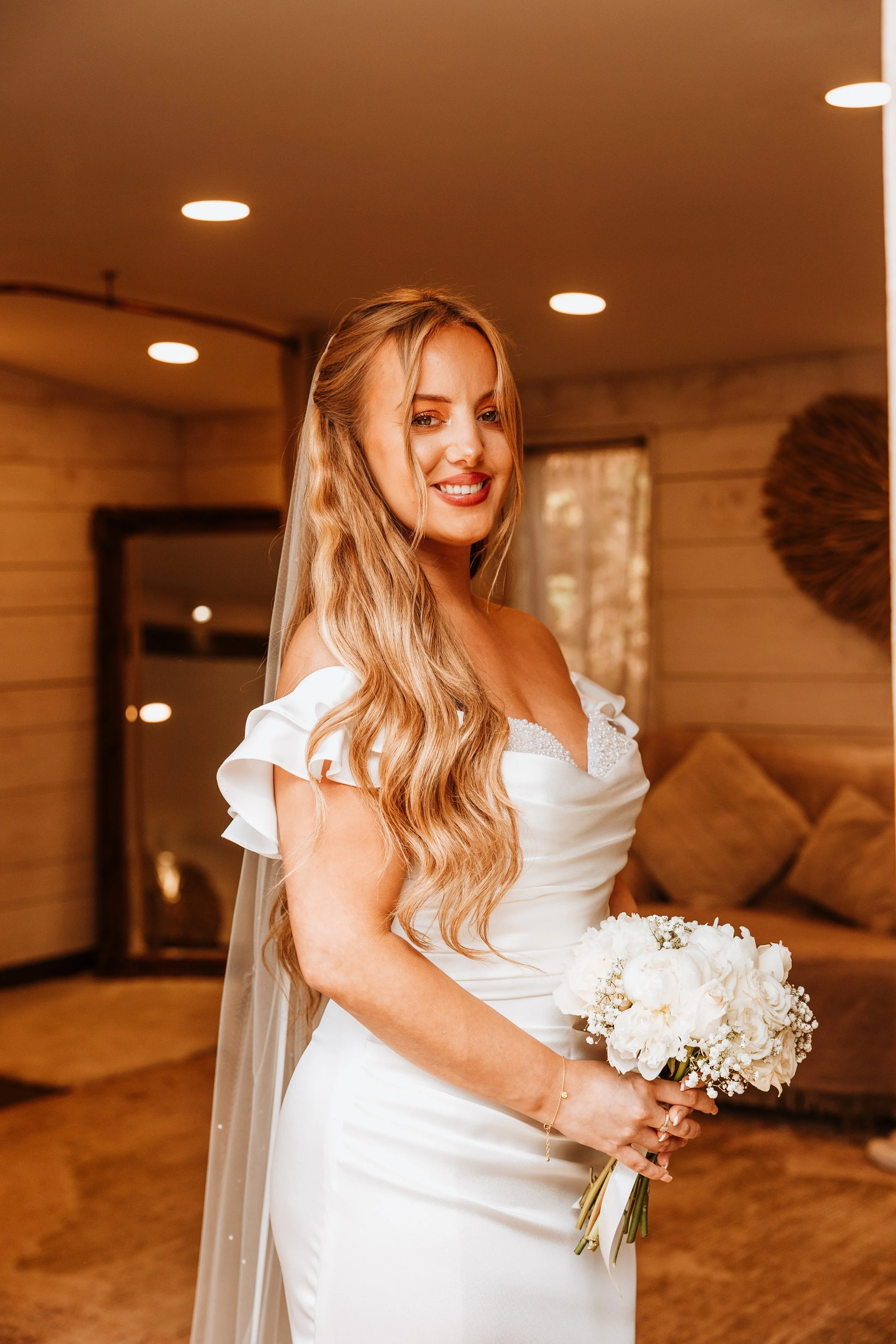 A bride in a white wedding dress holding a bouquet of white flowers, smiling in a warmly lit room with wood-paneled walls.