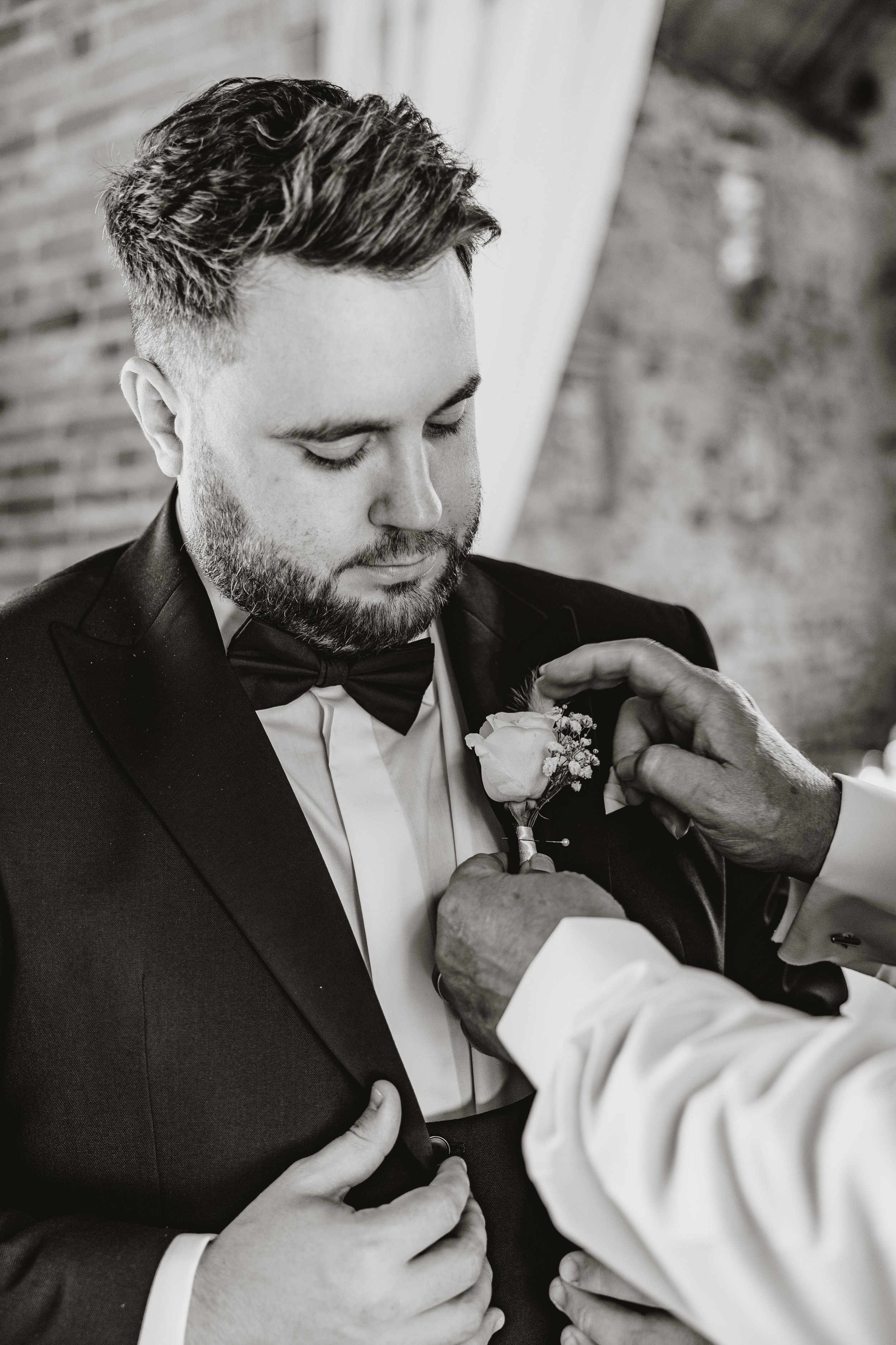 A man in a tuxedo with a bow tie is having a boutonniere pinned to his lapel by another person's hands.