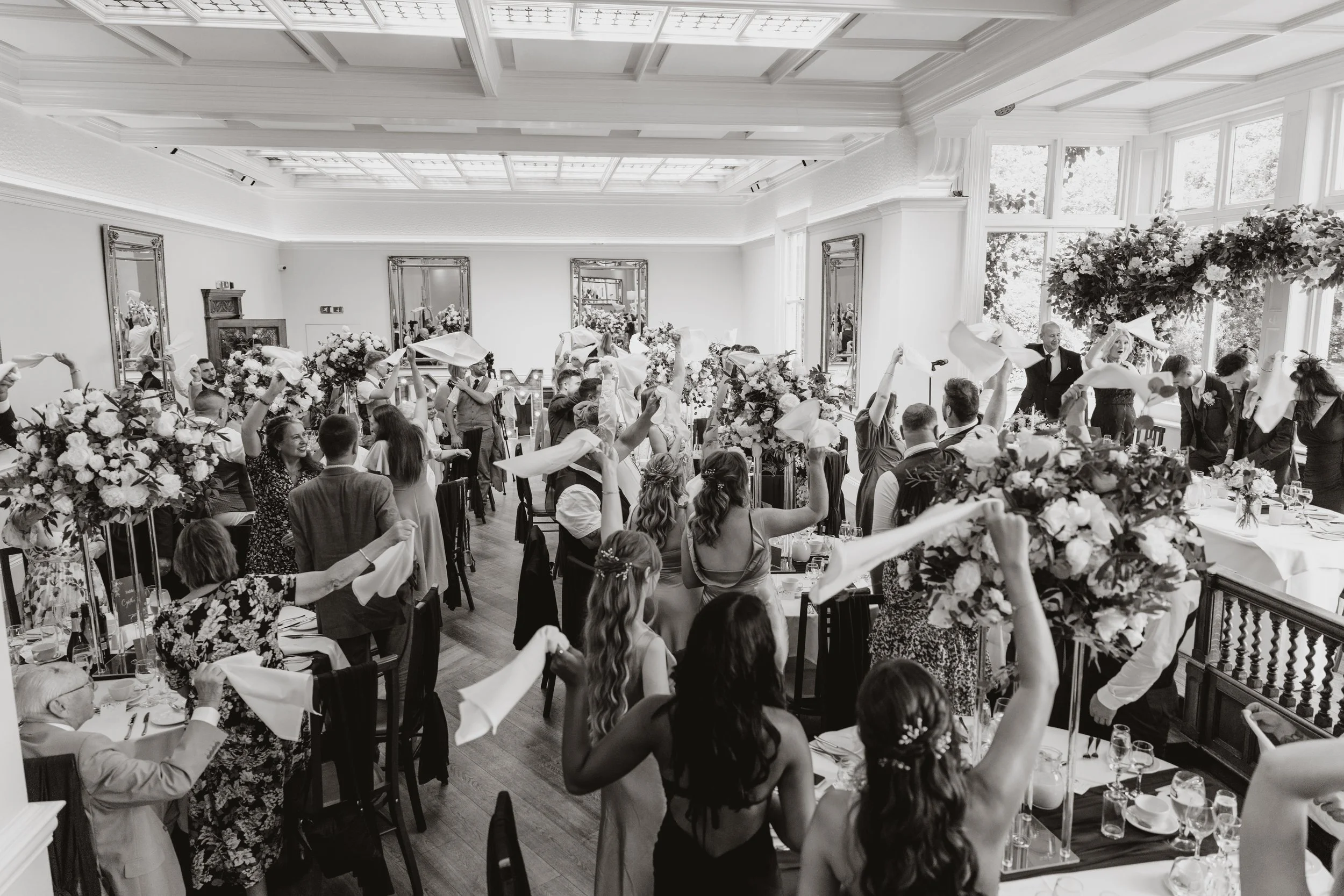 People celebrating at a wedding reception with waving napkins and large floral centerpieces in a bright, elegant room at Pendrall Hall, Codsall, West Midlands. 