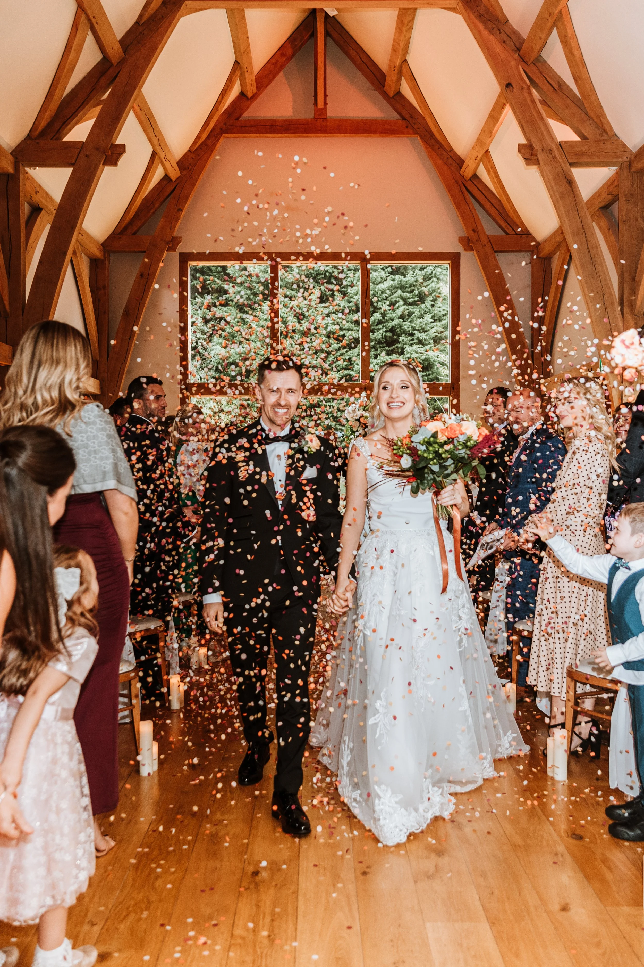 Happy bride and groom walking down the aisle, holding hands, with confetti falling around them inside a wooden chapel with large windows and guests celebrating.