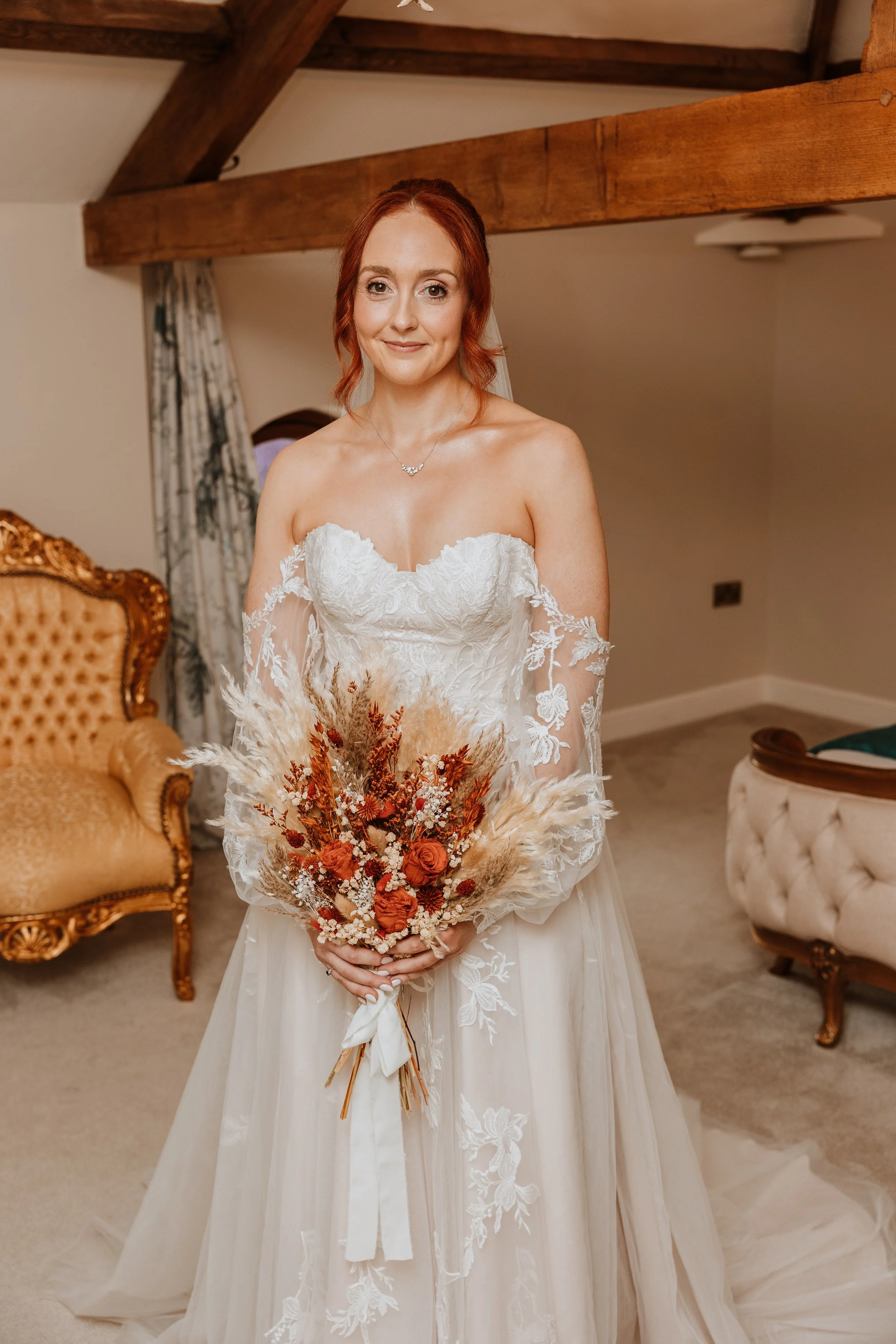 A bride with red hair and a strapless white lace wedding gown holding a bouquet of dried flowers in shades of orange, cream, and brown, standing in a cozy room with vintage furniture and wooden beams.