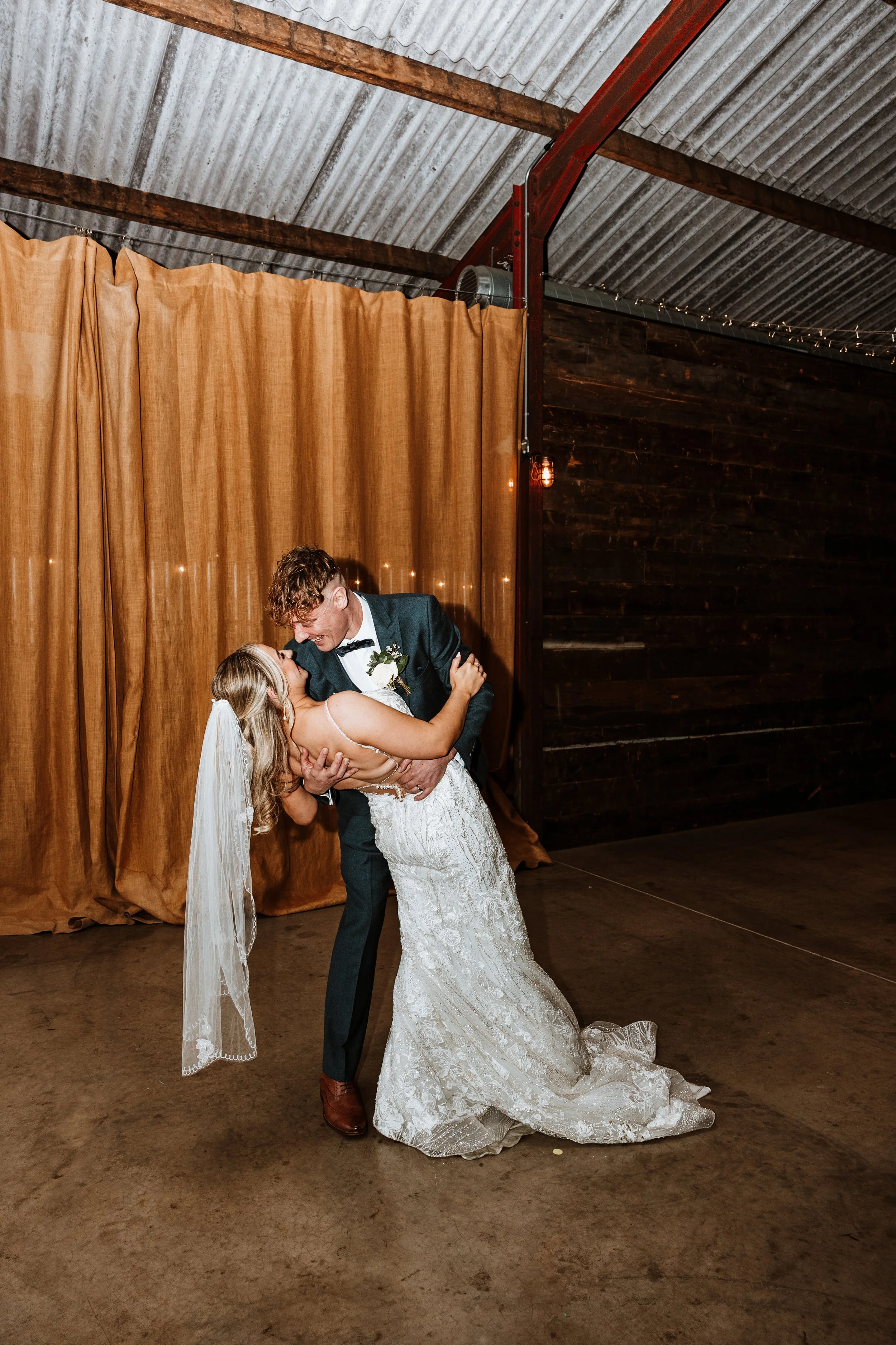 A newlywed couple sharing a dance, with the groom dipping the bride, in a rustic indoor wedding venue with warm lighting and a brown curtain backdrop.