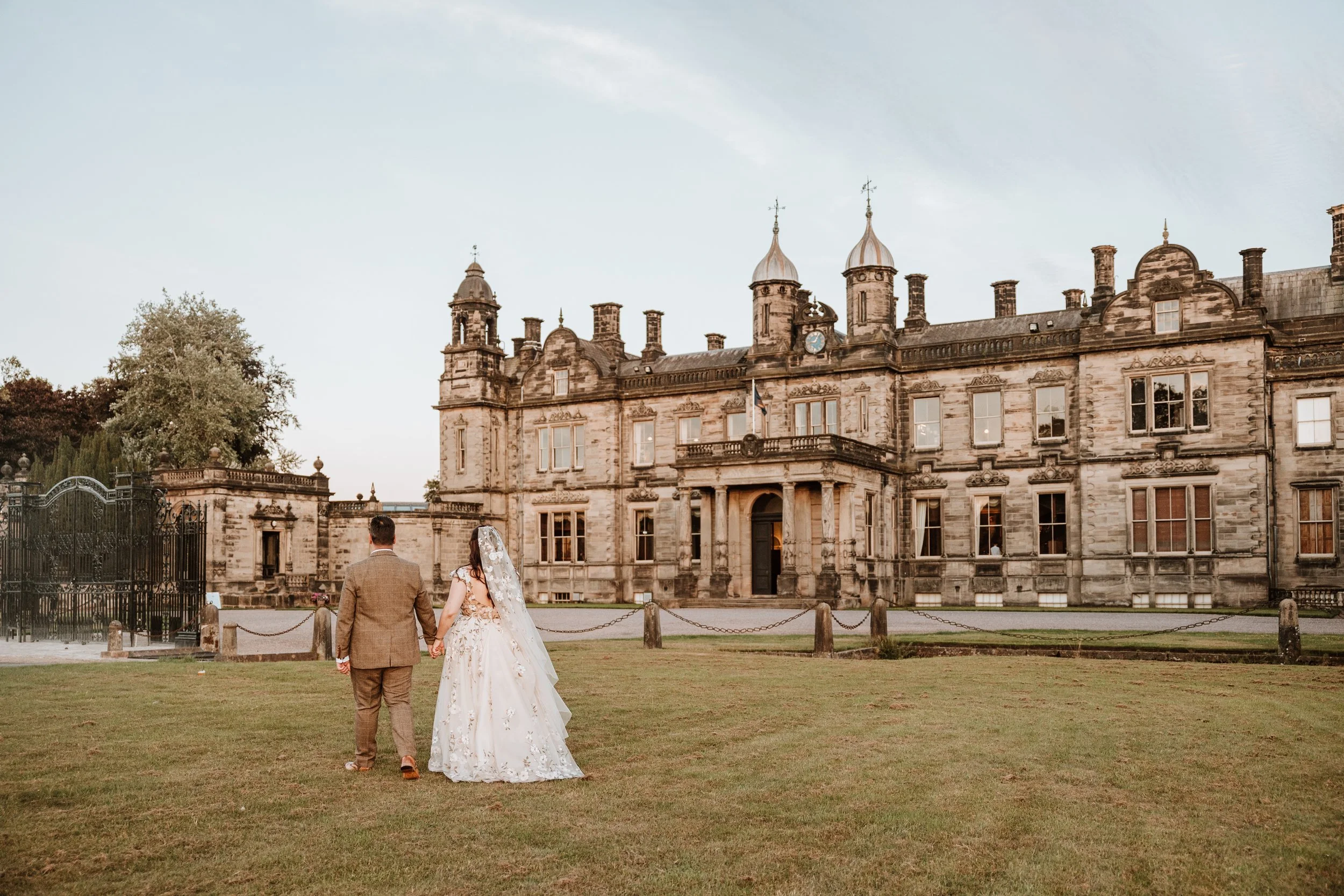 A bride and groom walking hand in hand on a grassy lawn towards a historic castle during sunset at Weston Hall, Staffordshire.