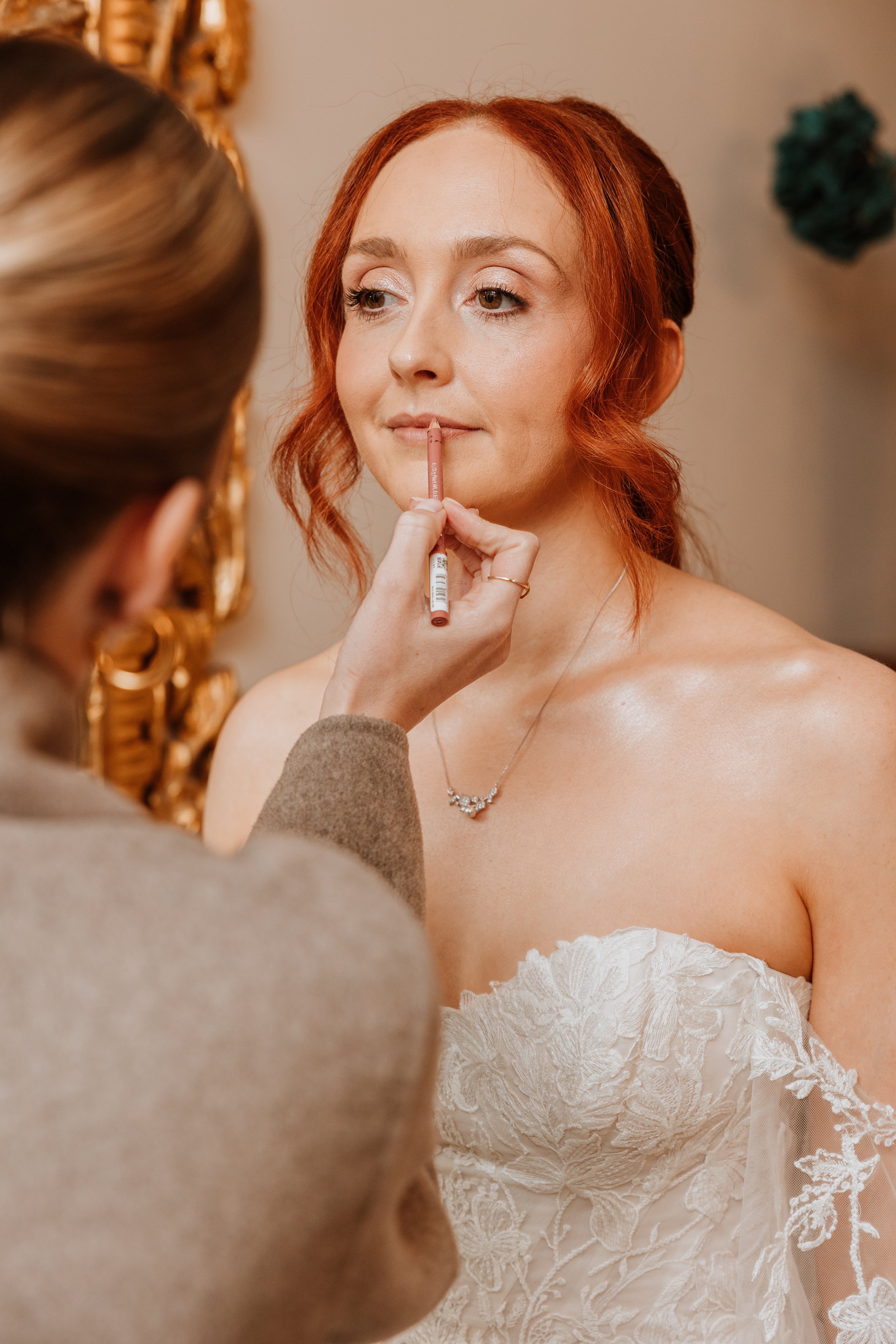 A woman with red hair getting her makeup done, wearing a white lace off-shoulder dress and a necklace, as a makeup artist applies lipstick.