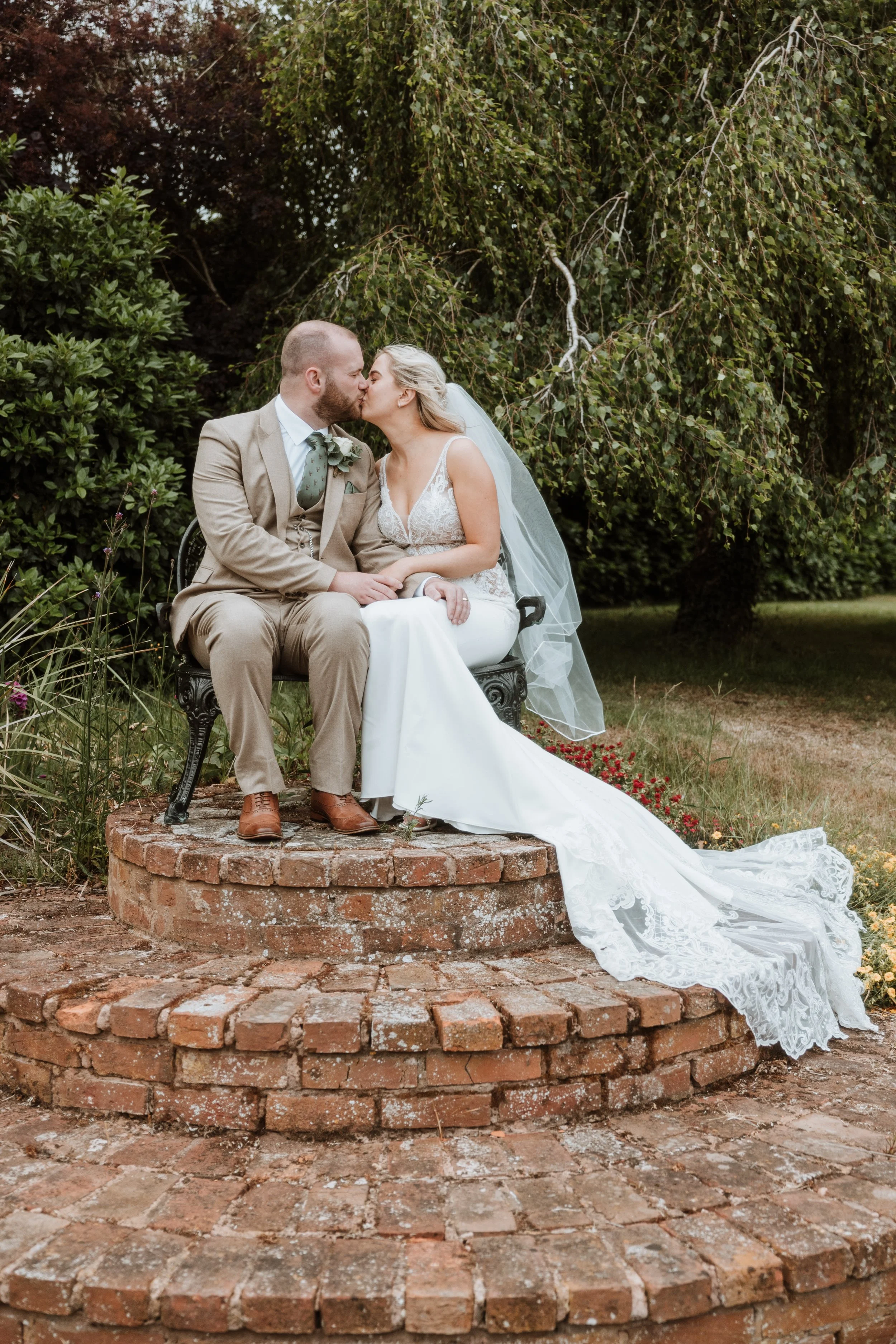 A bride and groom sitting on a black wrought iron bench on a brick platform, sharing a kiss outdoors. The bride is in a white wedding dress with lace details and a long train, and the groom is in a tan suit with a light green tie. There are green tre