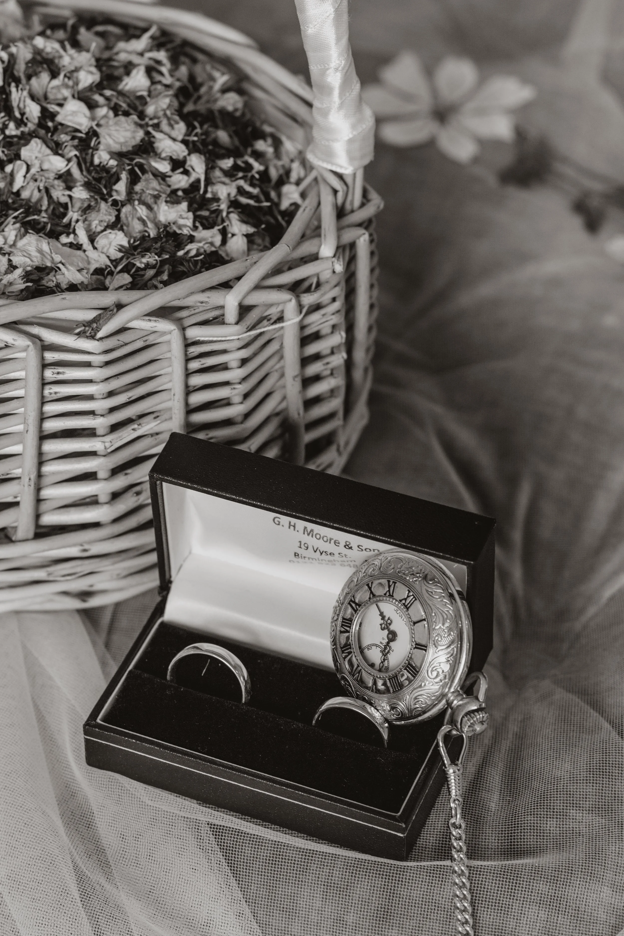 A vintage pocket watch with Roman numerals in a decorative case, placed inside a black presentation box with a chain attached, positioned on a textured surface next to a wicker basket filled with dried flowers.