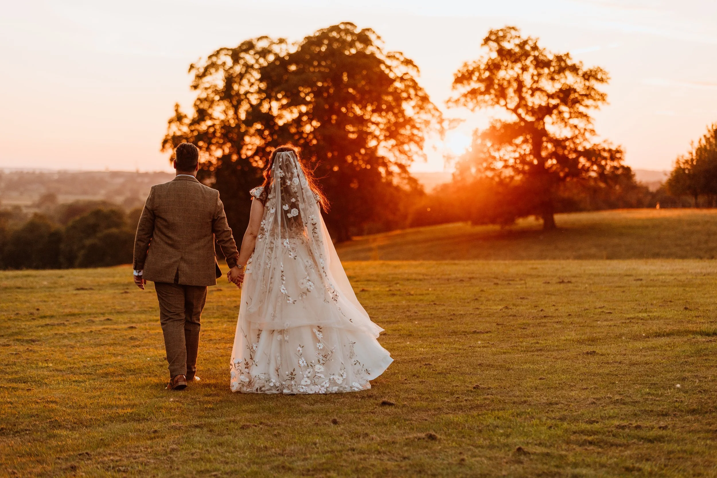A newlywed couple walking hand-in-hand across a grassy field at sunset, with trees and a sunset sky in the background.