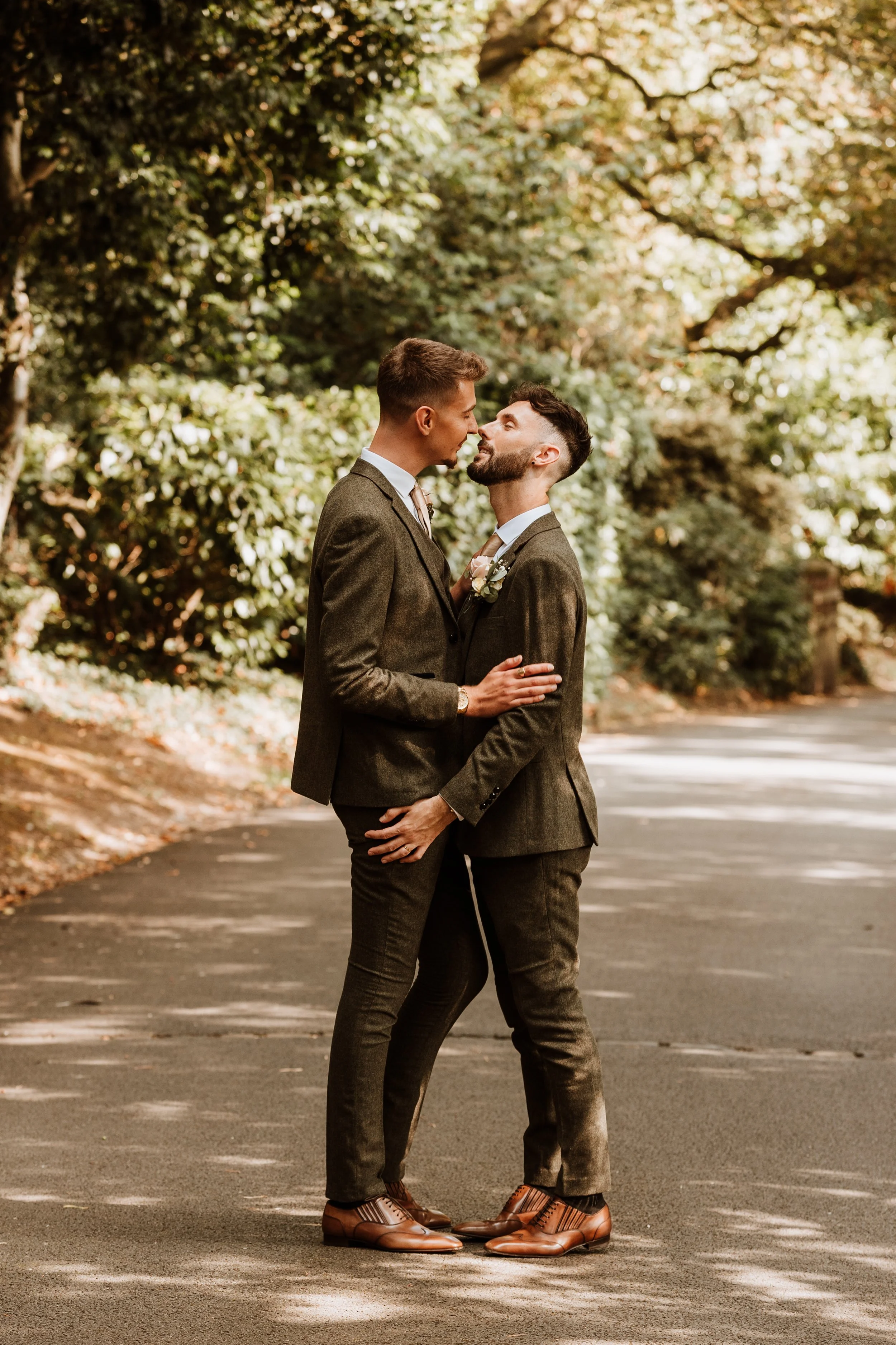 Two men in suits standing close together on a tree-lined street, about to kiss.