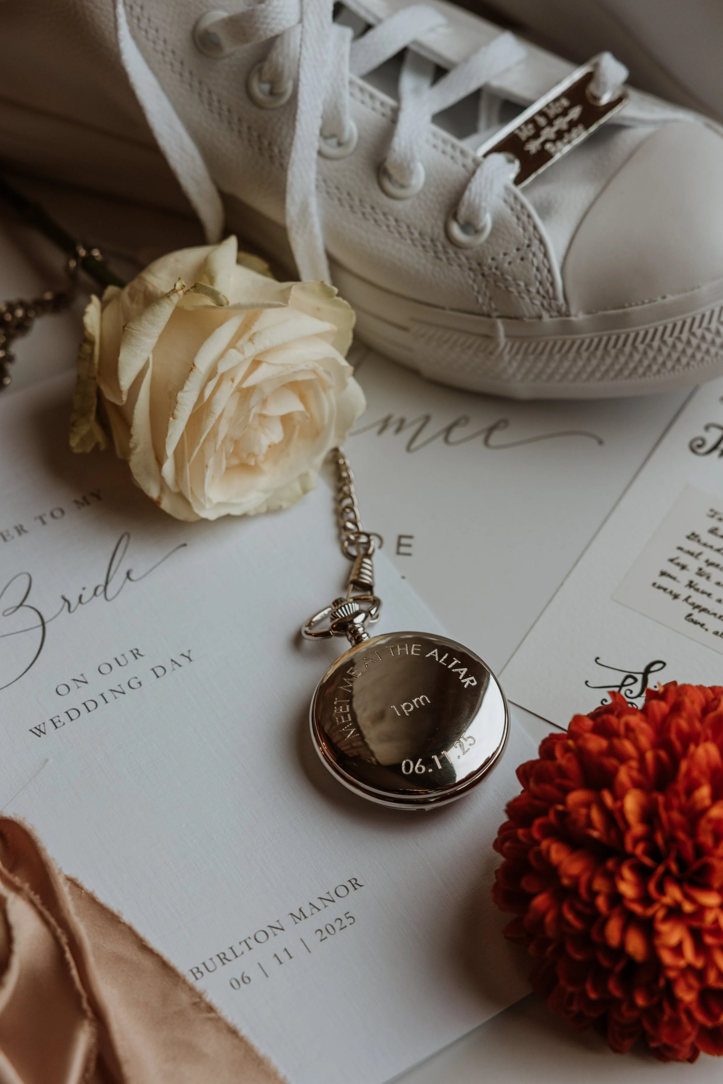A wedding invitation with wedding details, a white rose, a silver pocket watch engraved with the wedding date and location, a white sneaker, and an orange flower, arranged on a flat surface.