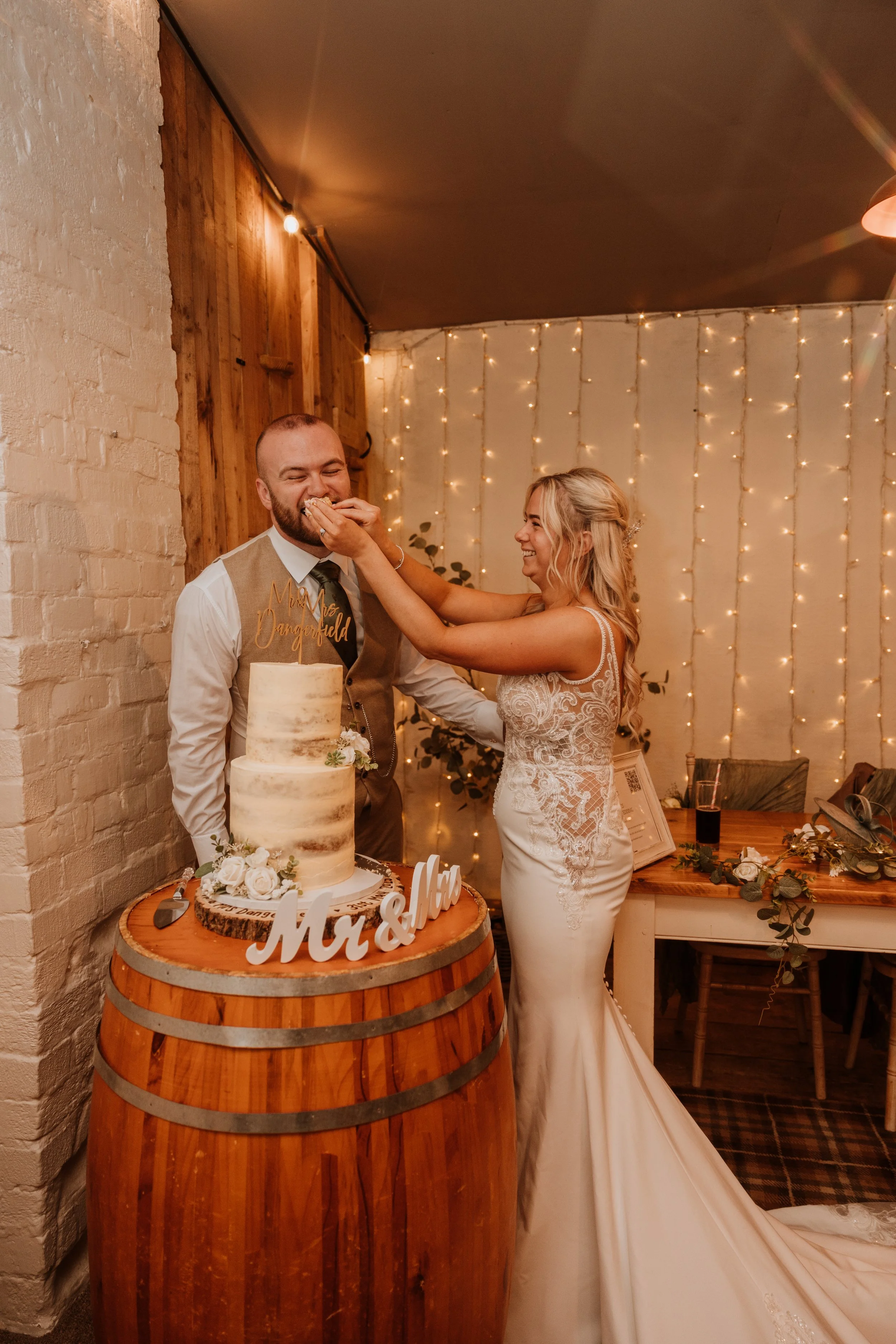 A bride and groom celebrating at their wedding reception. The bride is feeding cake to the groom while smiling, and they are standing behind a wedding cake on a wooden barrel. The background has string lights and wedding decor.