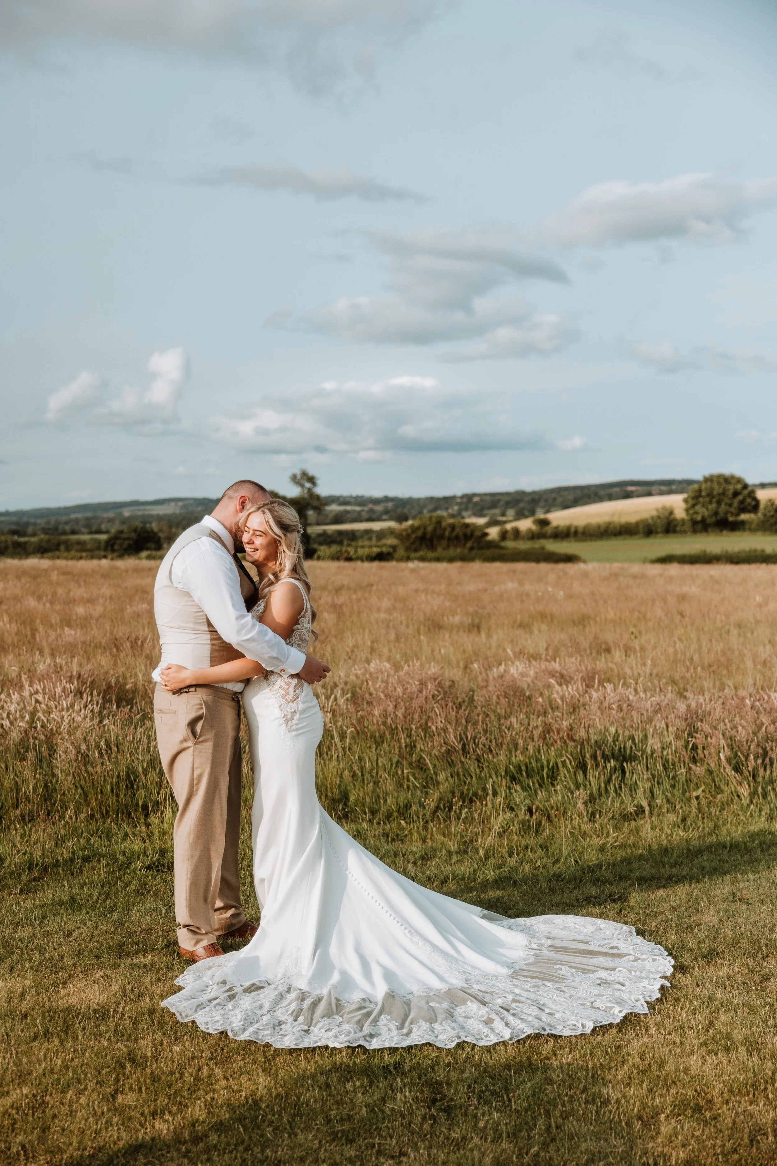 A bride and groom hugging outdoors in a grassy field with a scenic landscape and cloudy sky in the background.