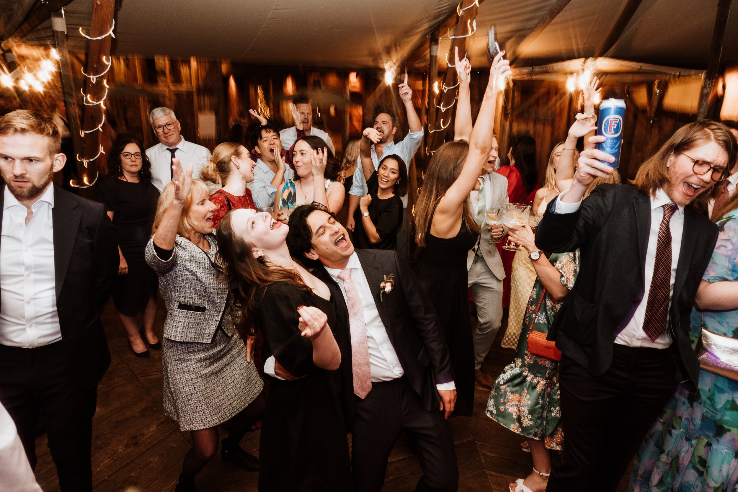 People dancing and celebrating at a wedding reception or party, some holding drinks, with everyone smiling and enjoying the event.