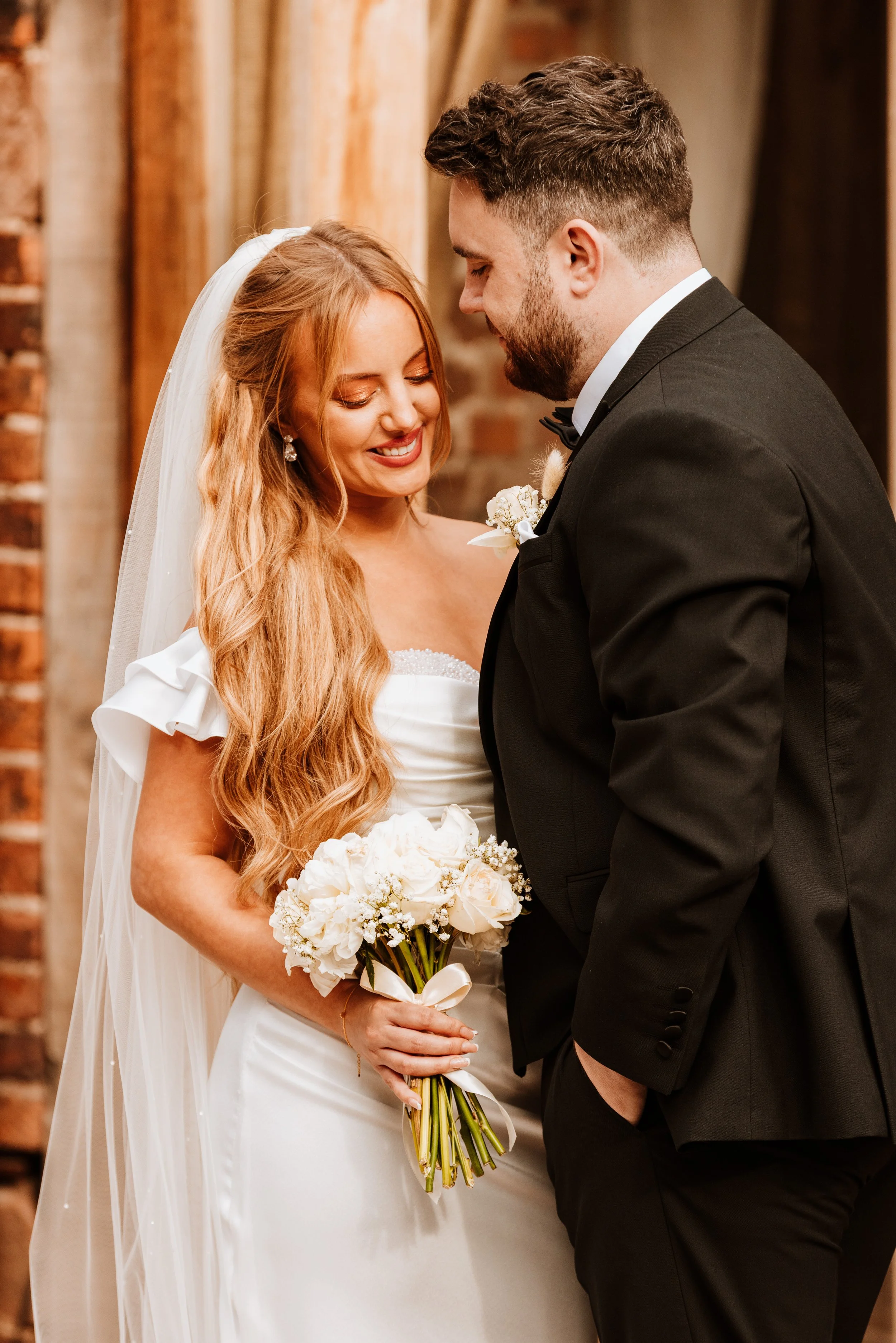A bride and groom sharing a tender moment at their wedding. The bride has long, wavy red hair, wearing a white wedding dress and holding a bouquet of white flowers. The groom has short dark hair and a beard, wearing a black tuxedo with a bow tie. The