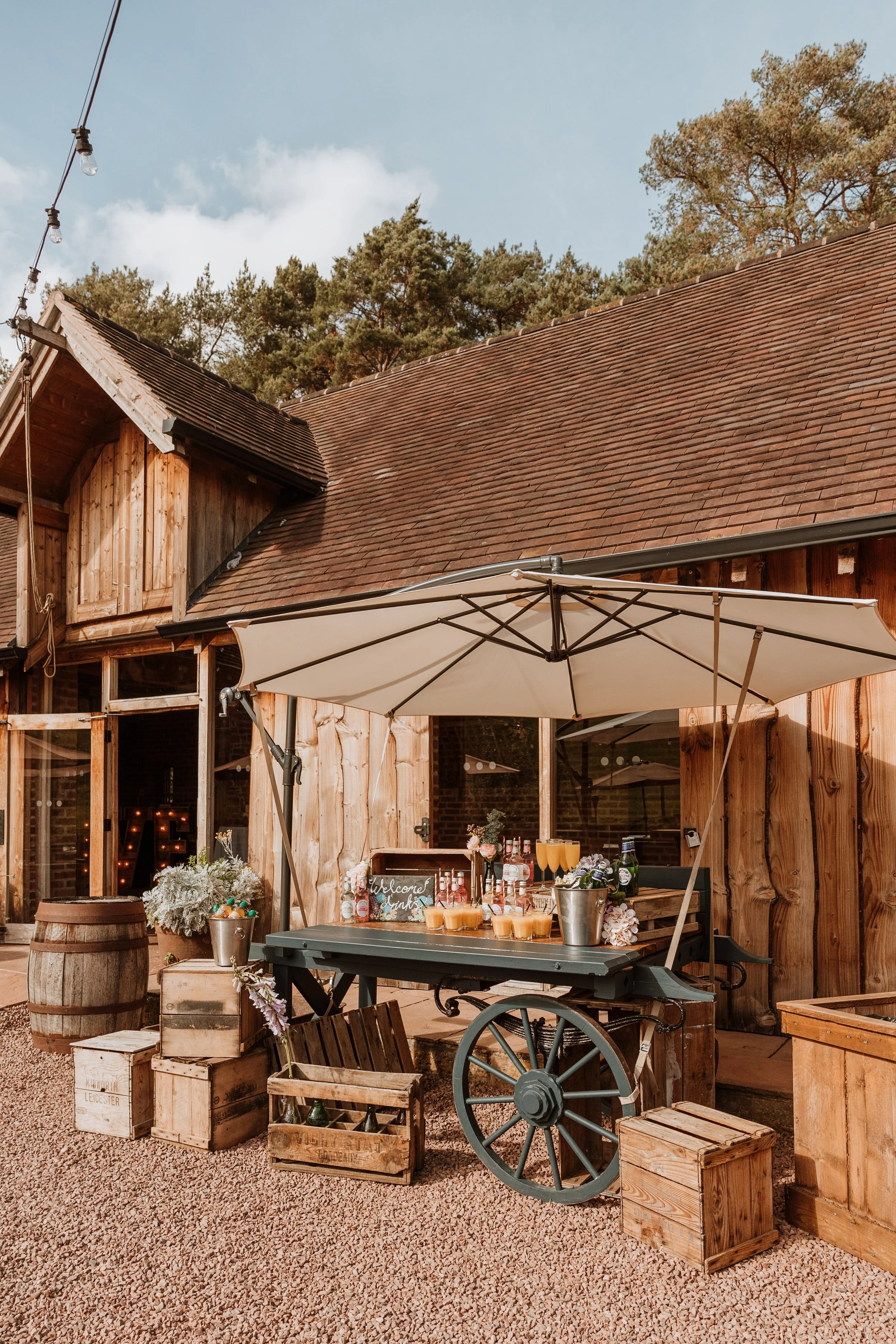 Outdoor rustic bar setup with a large white umbrella, wooden crates, a small wooden cart with drinks, and decorative flowers, in front of a wooden building with a sloped roof.