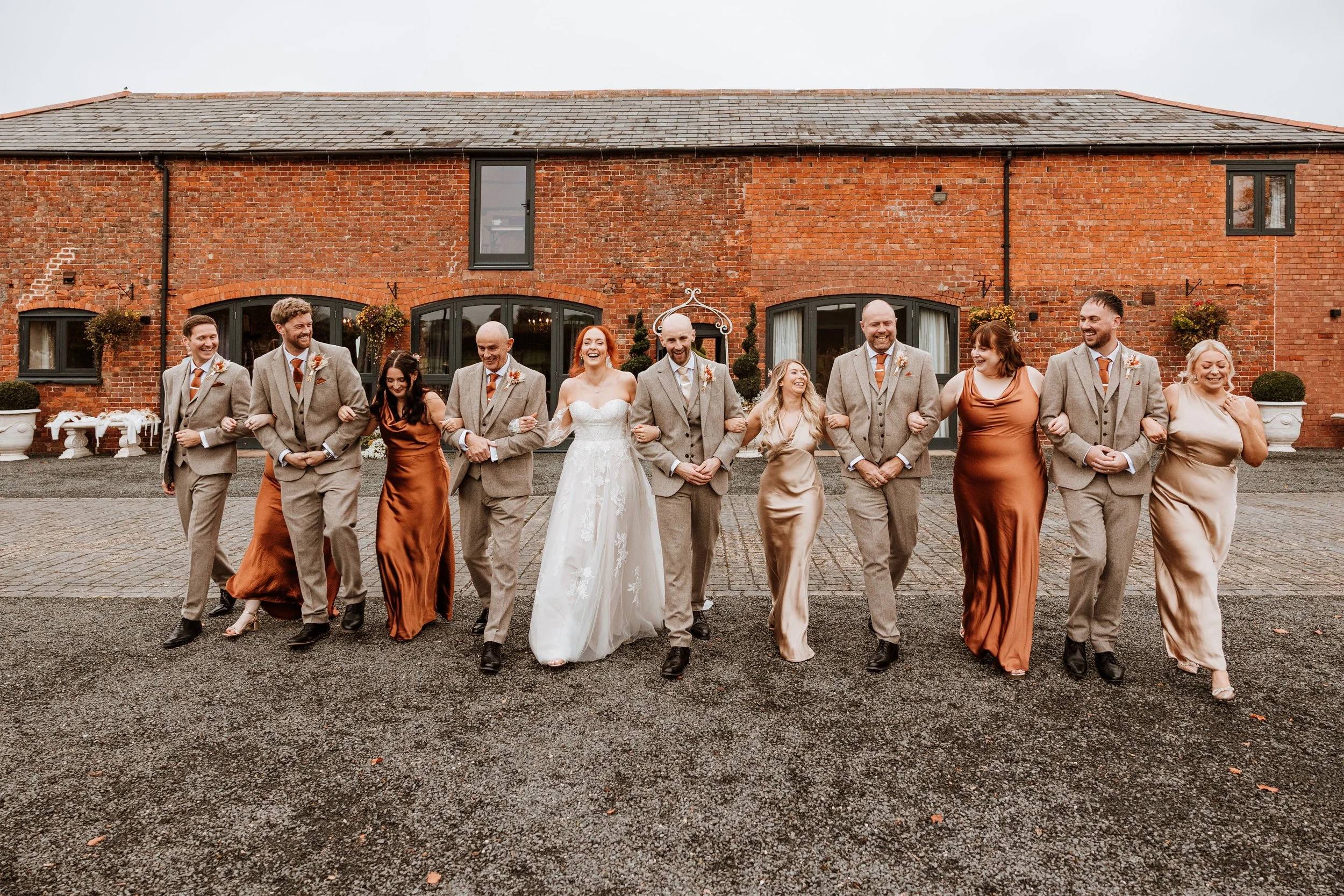 A wedding party of twelve people walking arm in arm outside a brick building. The bride is in the center wearing a white wedding dress, and the groom is to her right wearing a light gray suit with a beige vest. Bridesmaids are in satin dresses in sha