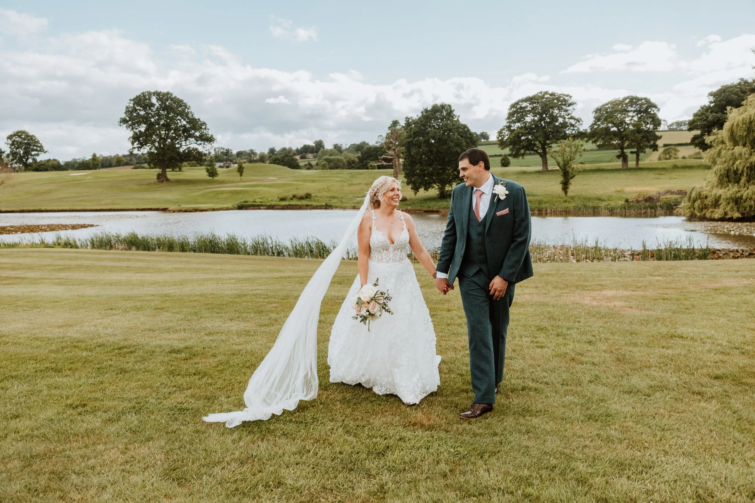A bride and groom holding hands and smiling at each other on a grassy area near a pond with trees and a cloudy sky in the background at The Astbury Hall and Golf Club, Bridgnorth.