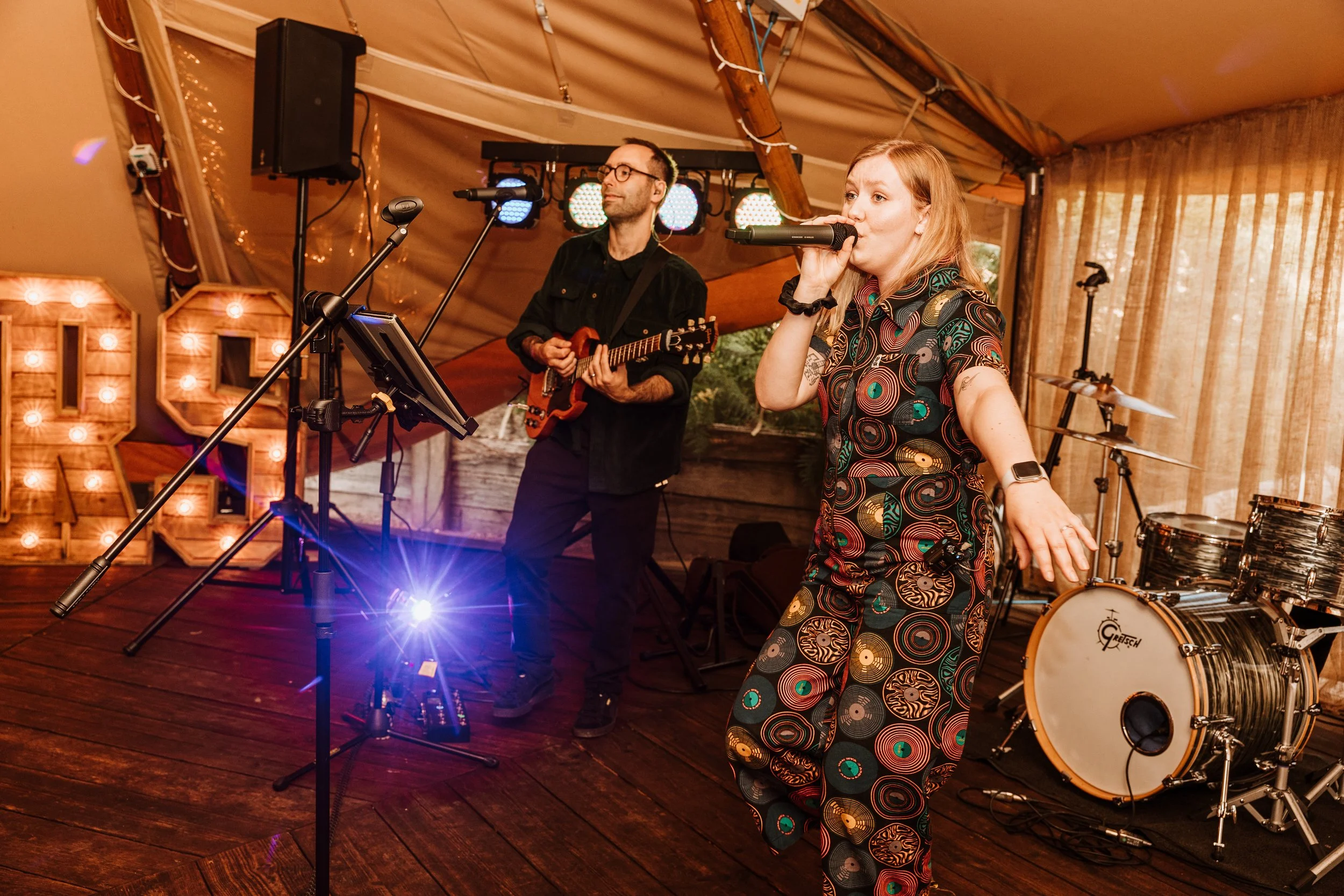 A woman singing into a microphone while a man plays electric guitar on a stage decorated with string lights and wooden letters.