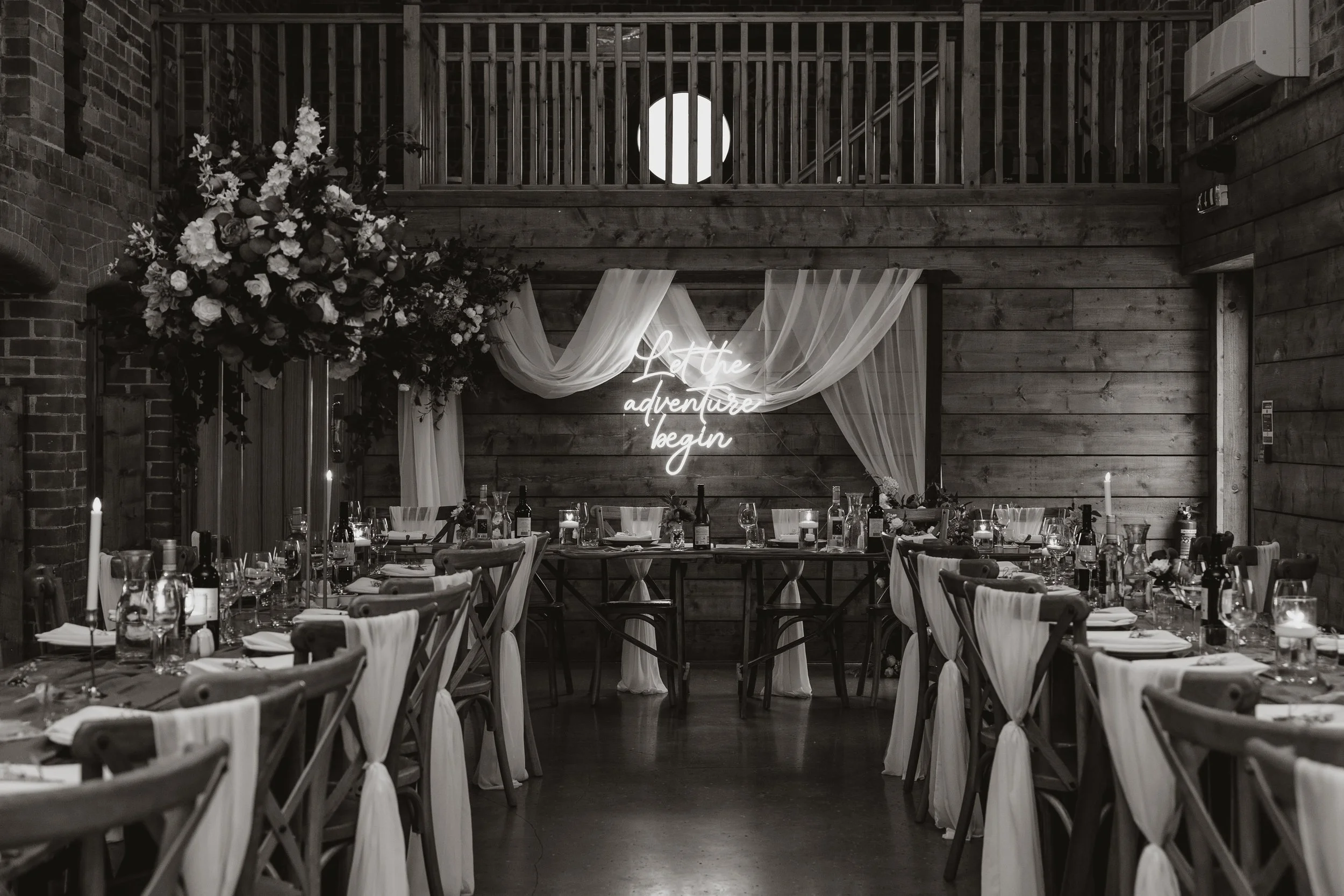 A decorated indoor dining area with a rustic wooden interior, featuring a table set with wine bottles, glasses, candles, and tableware. A floral arrangement and a white fabric backdrop with a neon sign reading "Let the adventure begin" are also visib