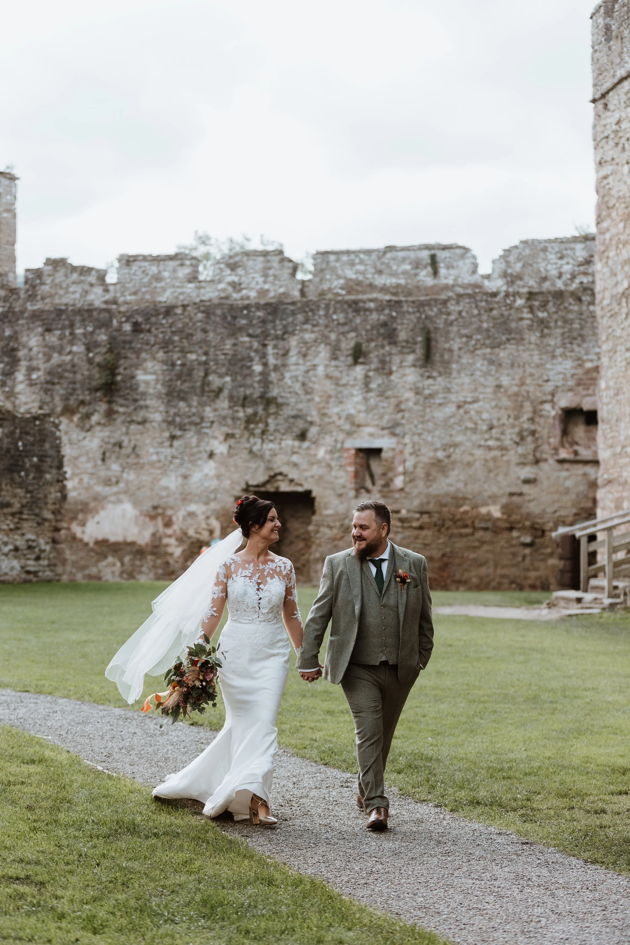 A bride and groom walking hand in hand on a gravel path outside, with a historic stone castle in the background during daytime.