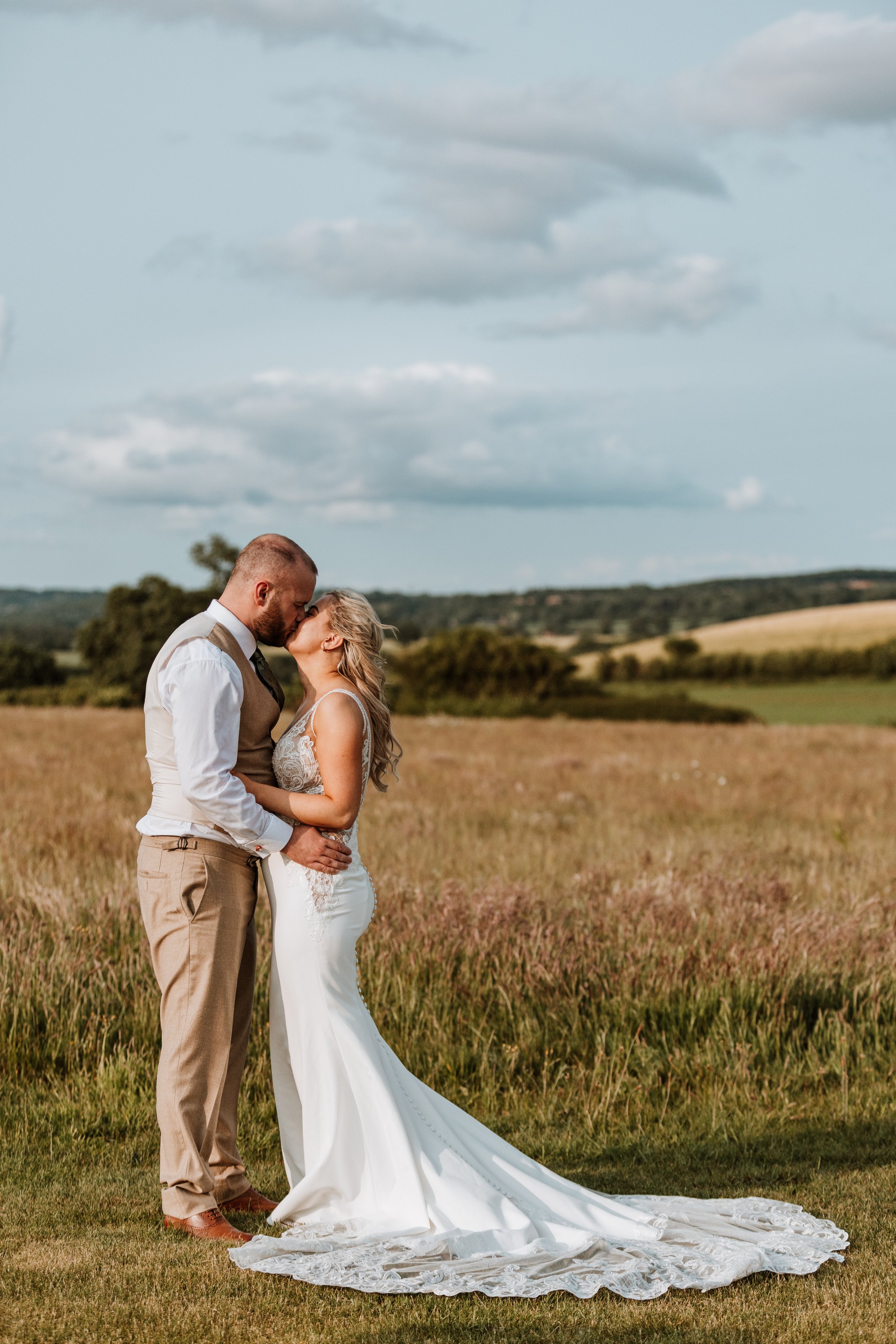 A couple in wedding attire sharing a kiss in a field with hills and clouds in the background.