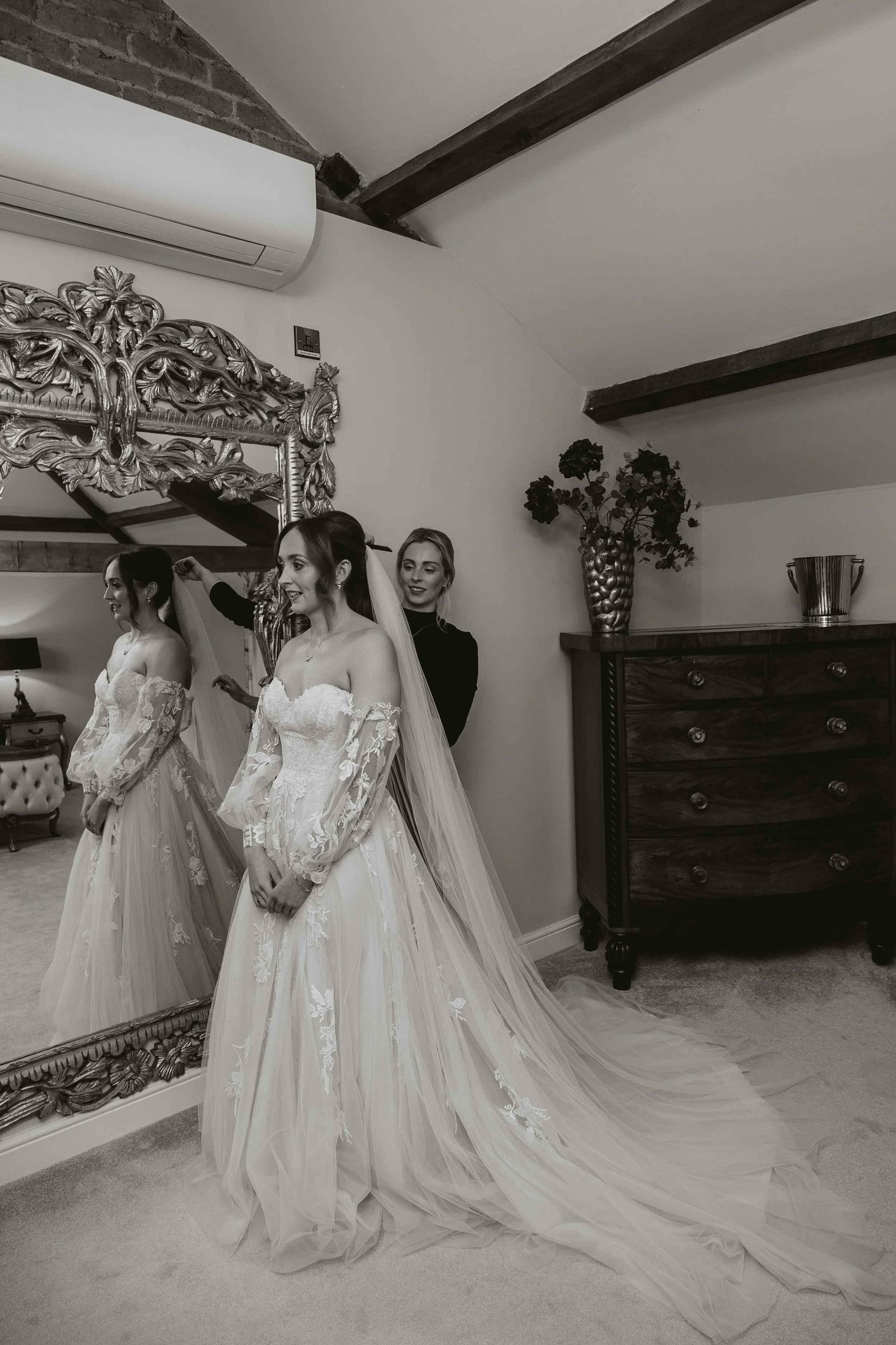 A bride in a lace wedding gown with long sleeves and a long train stands in front of a mirror, while a woman helps her adjust her veil in a bedroom with a wooden dresser, a large ornate mirror, and decorative objects.