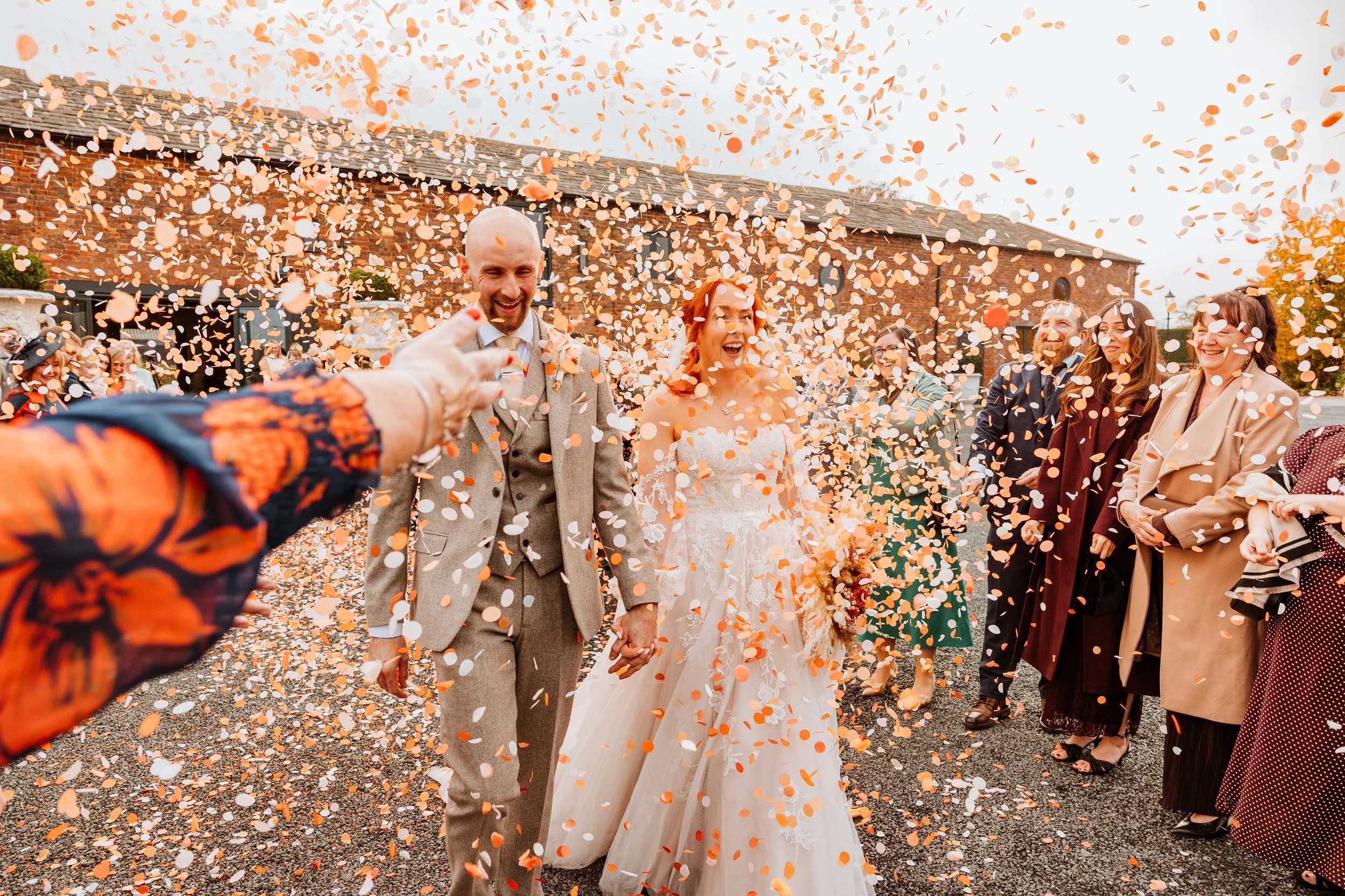 Bride and groom holding hands smiling as guests throw orange and white confetti during outdoor wedding celebration.