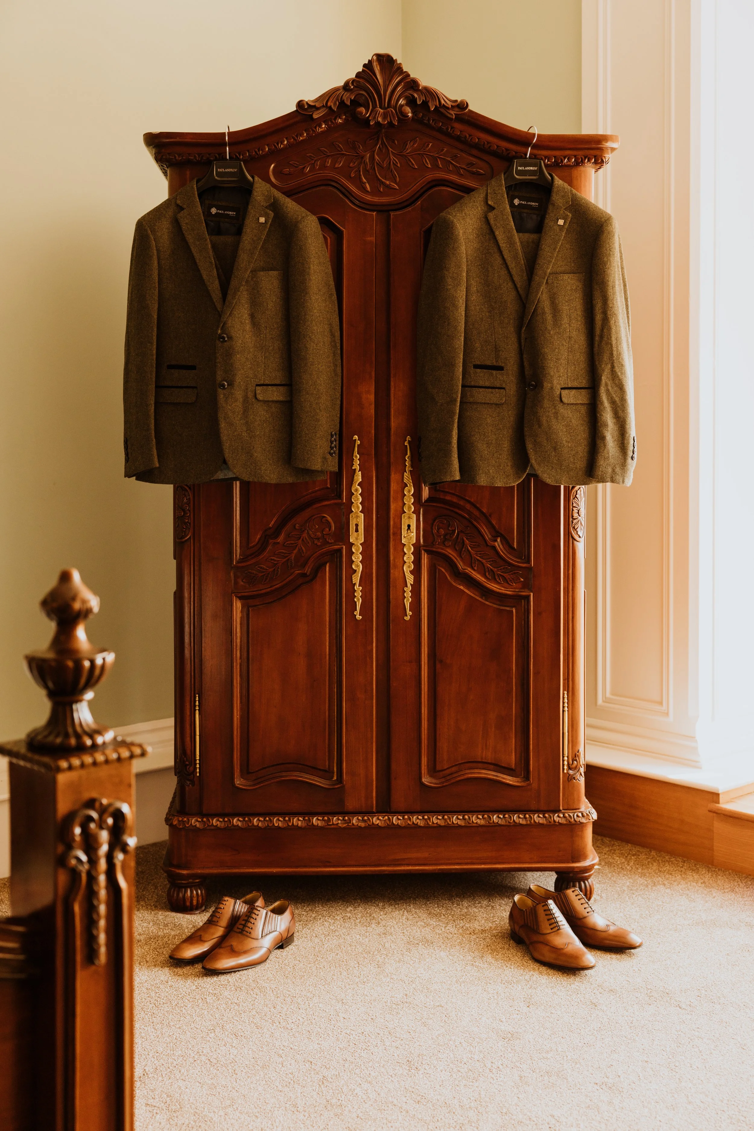 Two brown suits hanging on a wooden wardrobe, with matching brown shoes on the floor underneath.