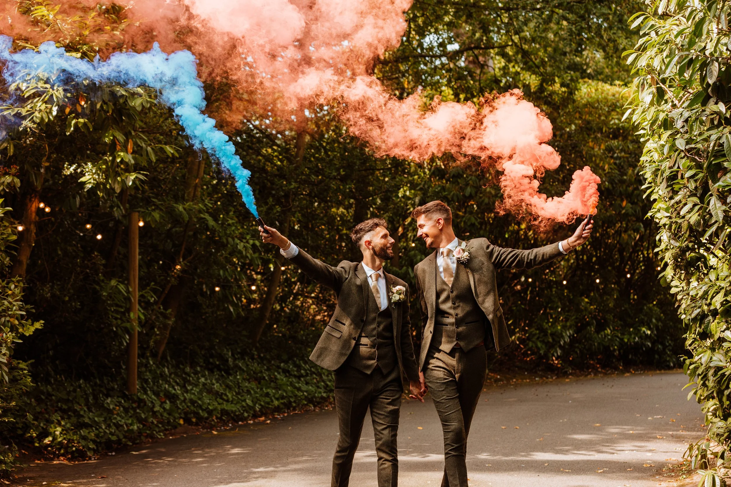 Same sex male couple in wedding suits holding colourful smoke bombs and walking hand in hand outdoors in a wooded area with string lights. They are smiling at each other.