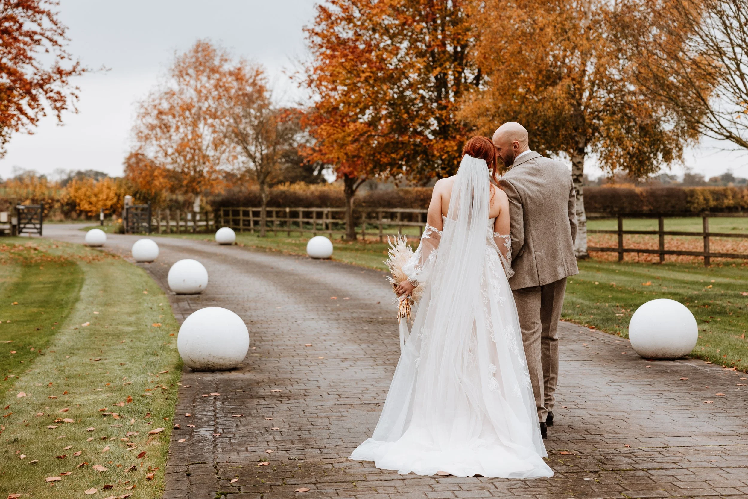 Bride and groom walking on a brick path outdoors during autumn, surrounded by trees with orange and red leaves, with large white spherical decorations along the side.