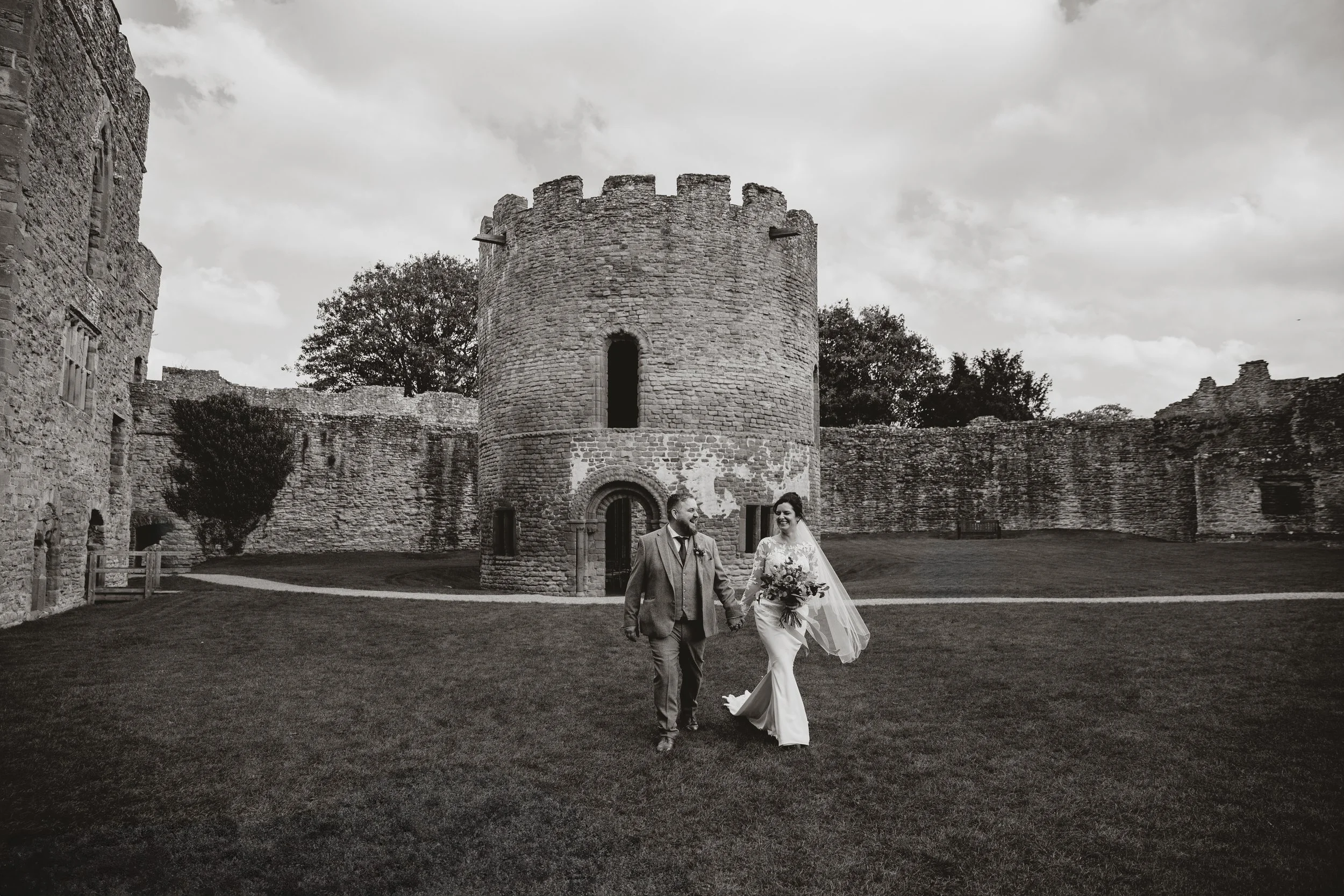 Black and white photo of a newlywed couple walking hand in hand in front of an old castle with a round tower and stone walls, cloudy sky background at Ludlow Castle.
