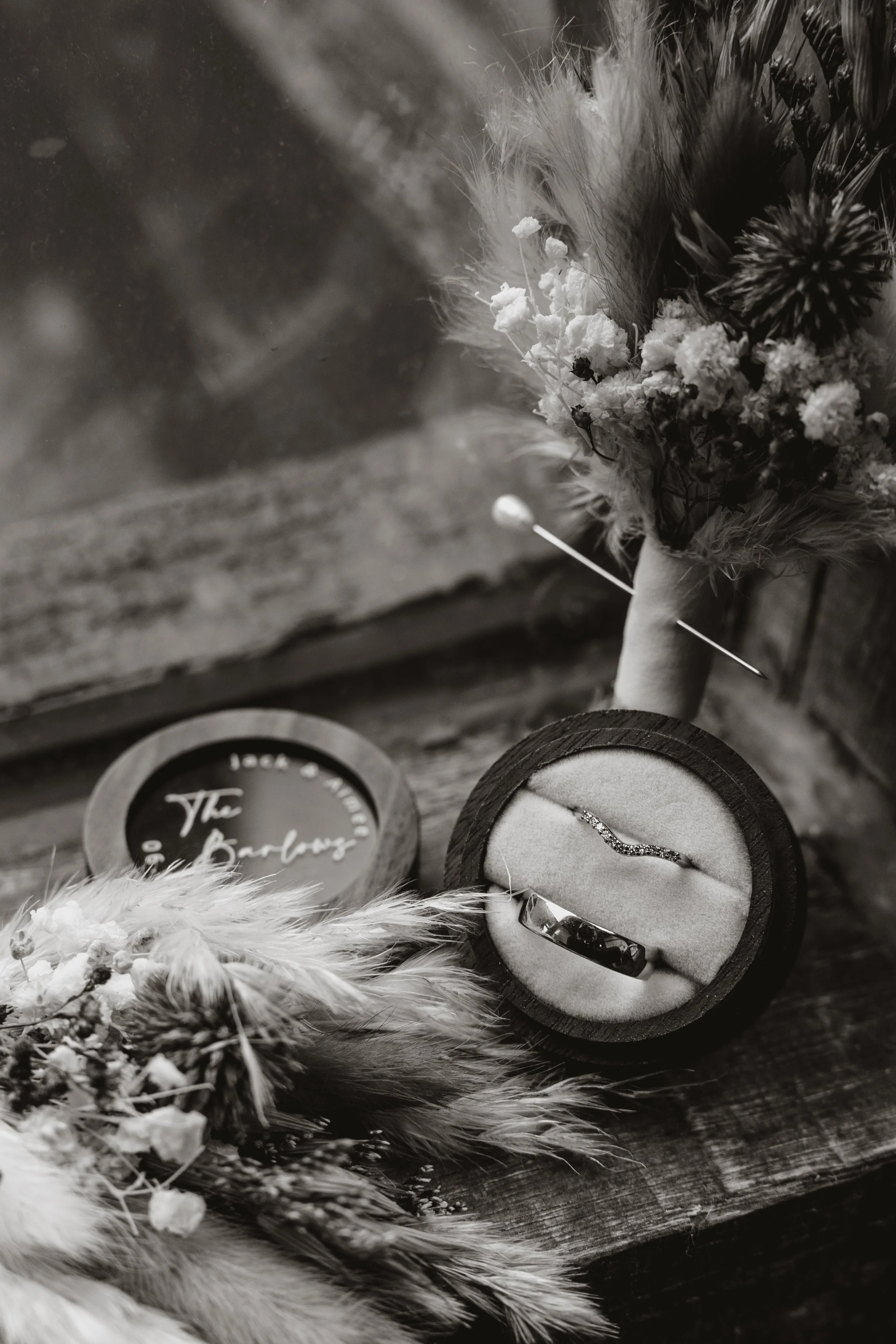 A black-and-white photo of a jewelry box with a wedding ring and matching ring, floral bouquets, and a framed sign stating 'The Brides' on a rustic wooden surface.
