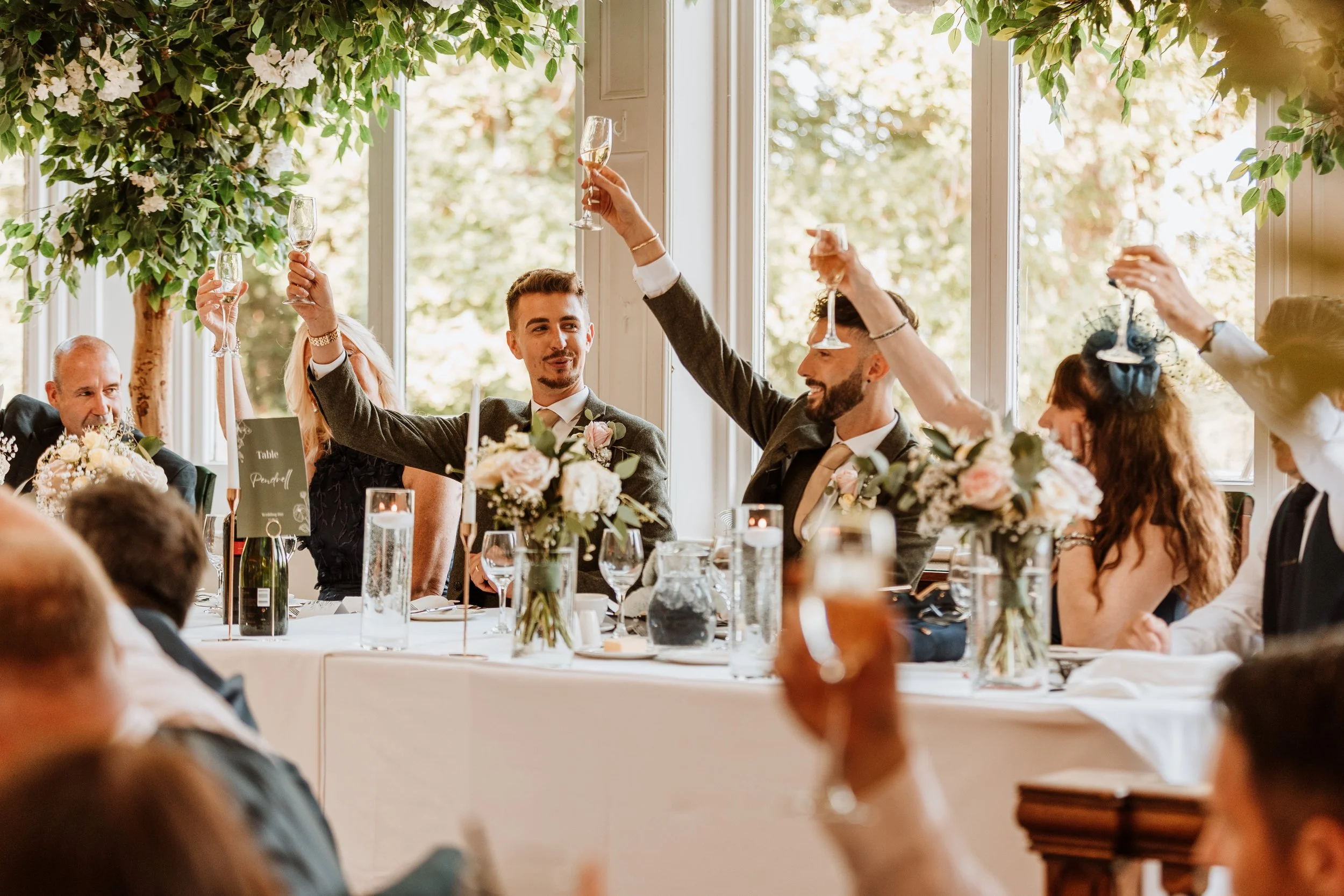 People raising glasses at a wedding reception, sitting at a decorated table with flowers and candles, with large windows and greenery outside in the background.