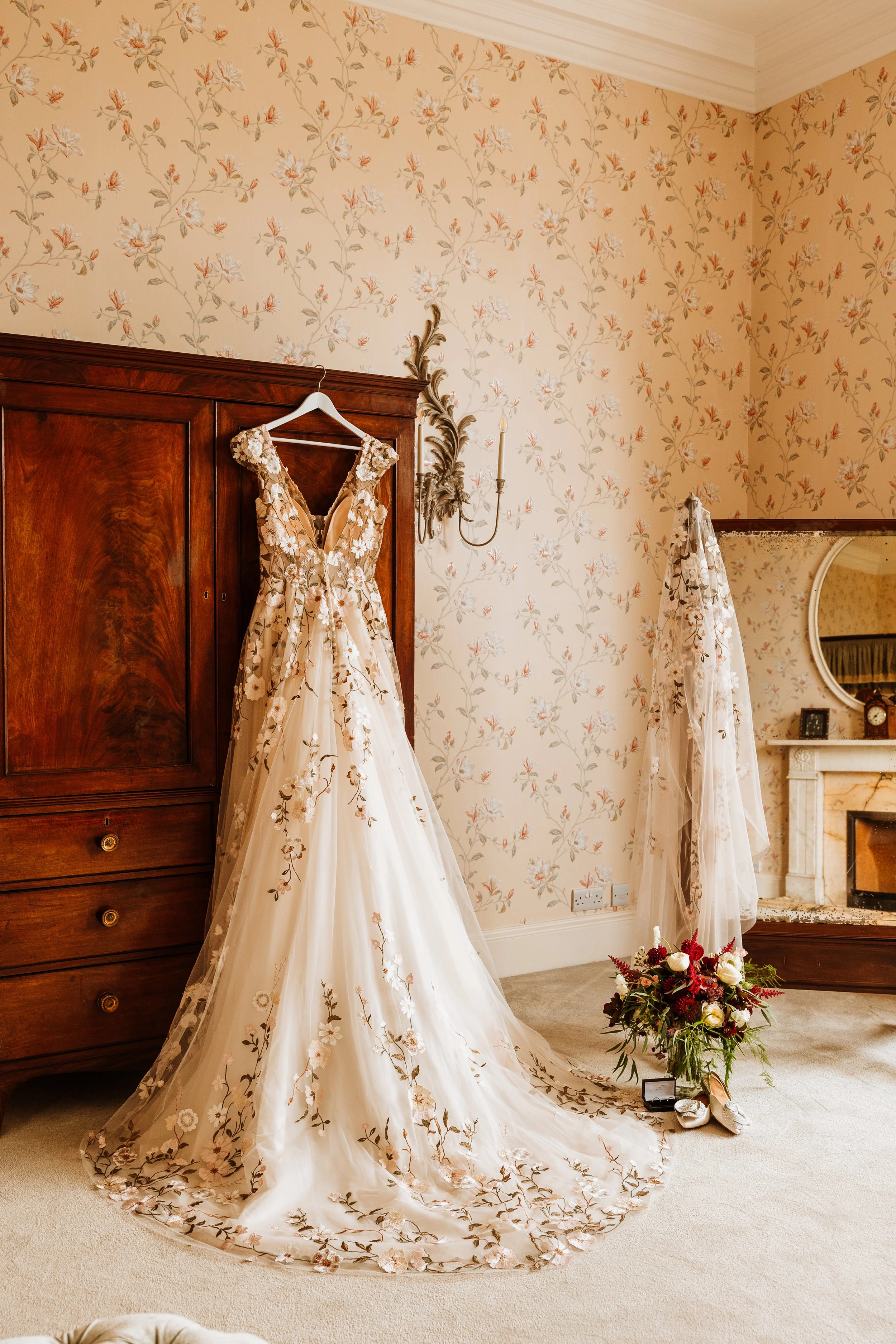 An elegant wedding dress with floral embroidery hanging on a wardrobe in a vintage-style room with floral wallpaper and a bouquet of flowers on the floor at Weston Hall, Staffordshire.