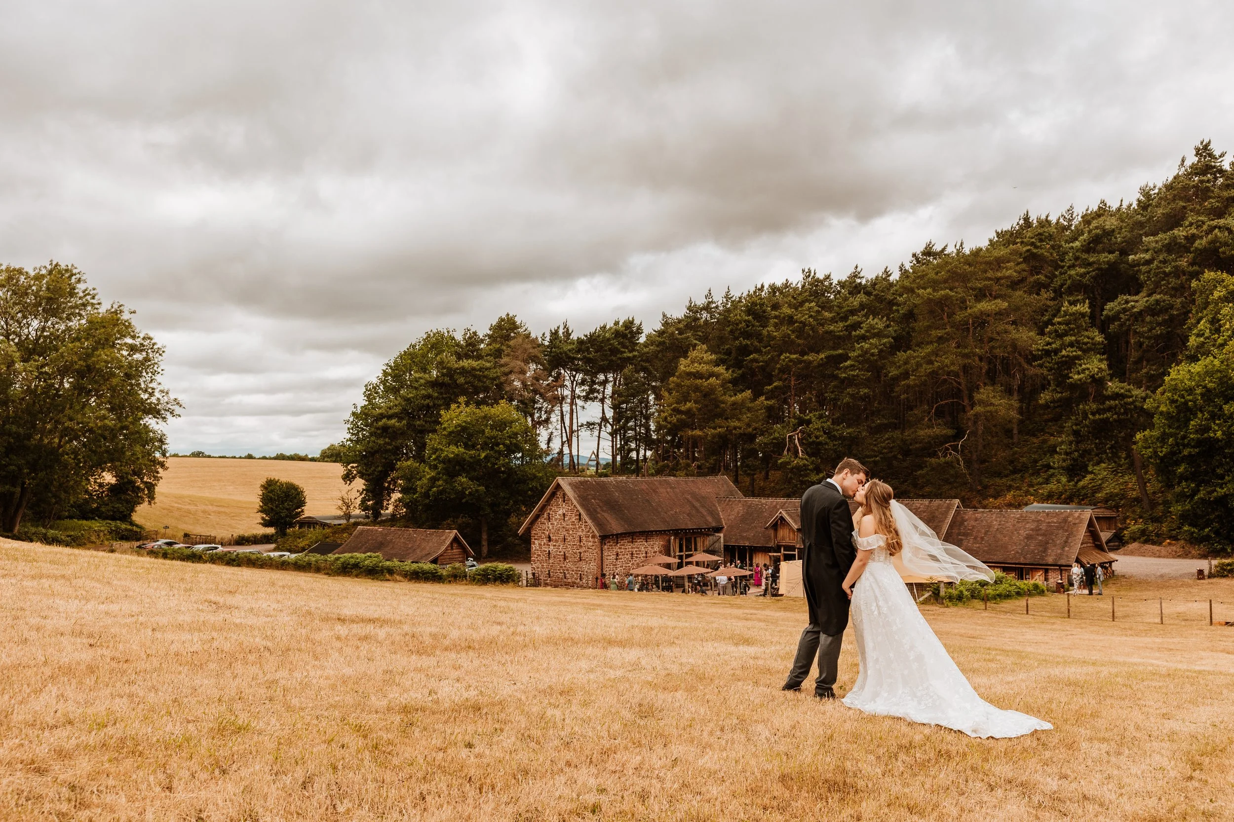 A bride and groom sharing a kiss on a grassy field outside a rustic barn, with a backdrop of trees and cloudy sky.