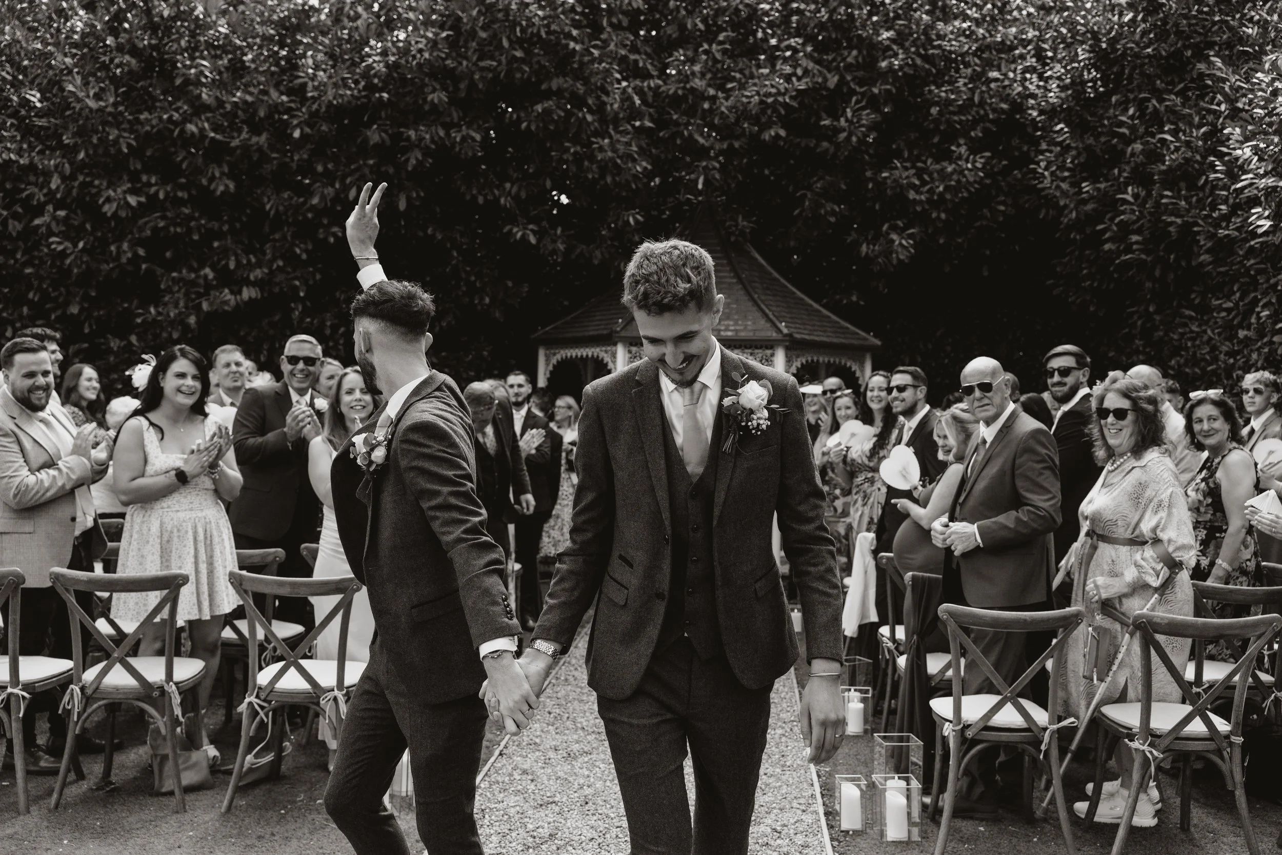 A gay couple holding hands and walking down the aisle after their wedding ceremony, surrounded by smiling guests at an outdoor wedding.
