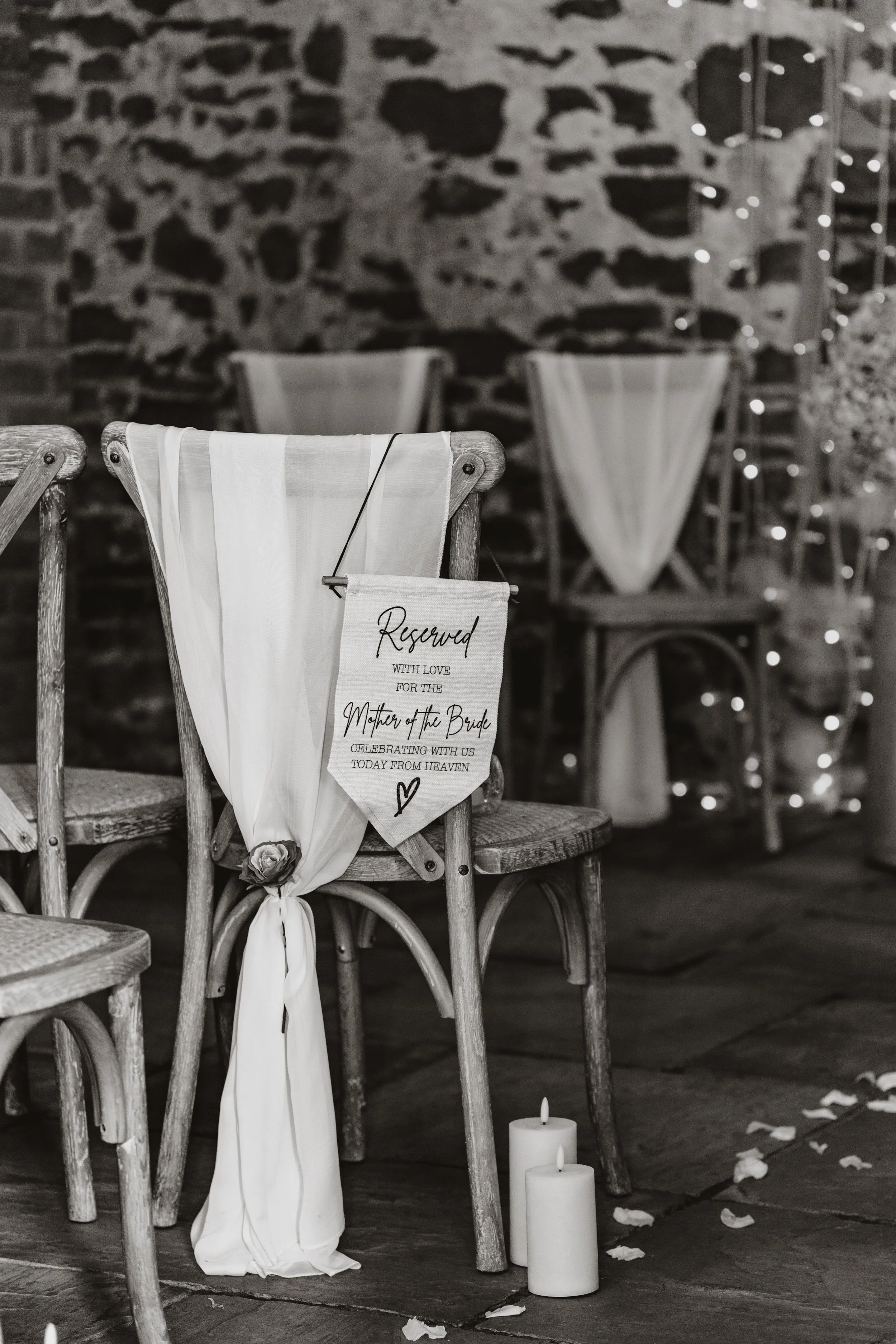 Wedding ceremony reserved sign hanging on a wooden chair draped with white cloth, with candles and flower petals on the floor, a rustic brick wall in the background.