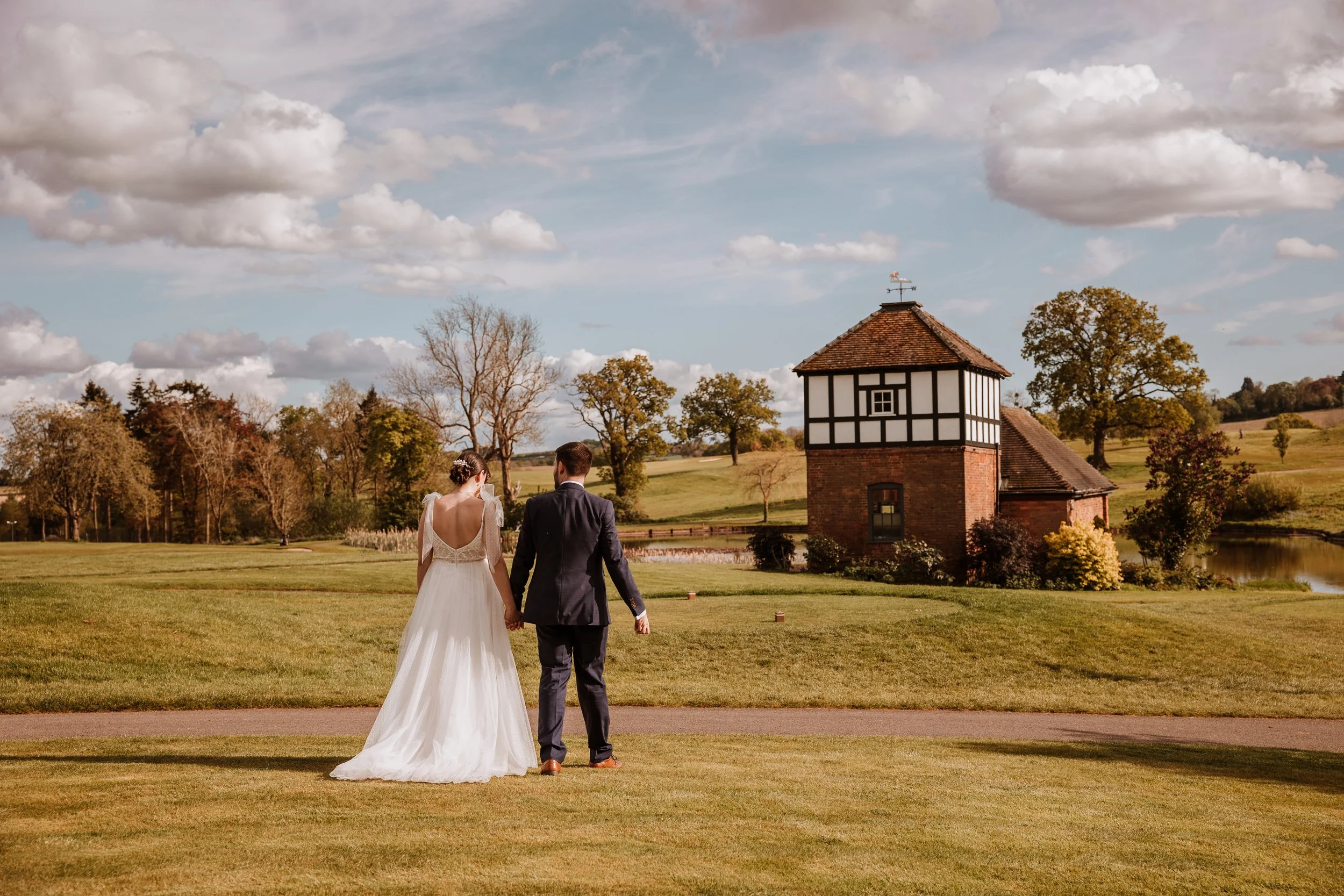 A bride and groom walking hand in hand on a grassy field near a small pond, with a historic timber-framed building under a partly cloudy sky.