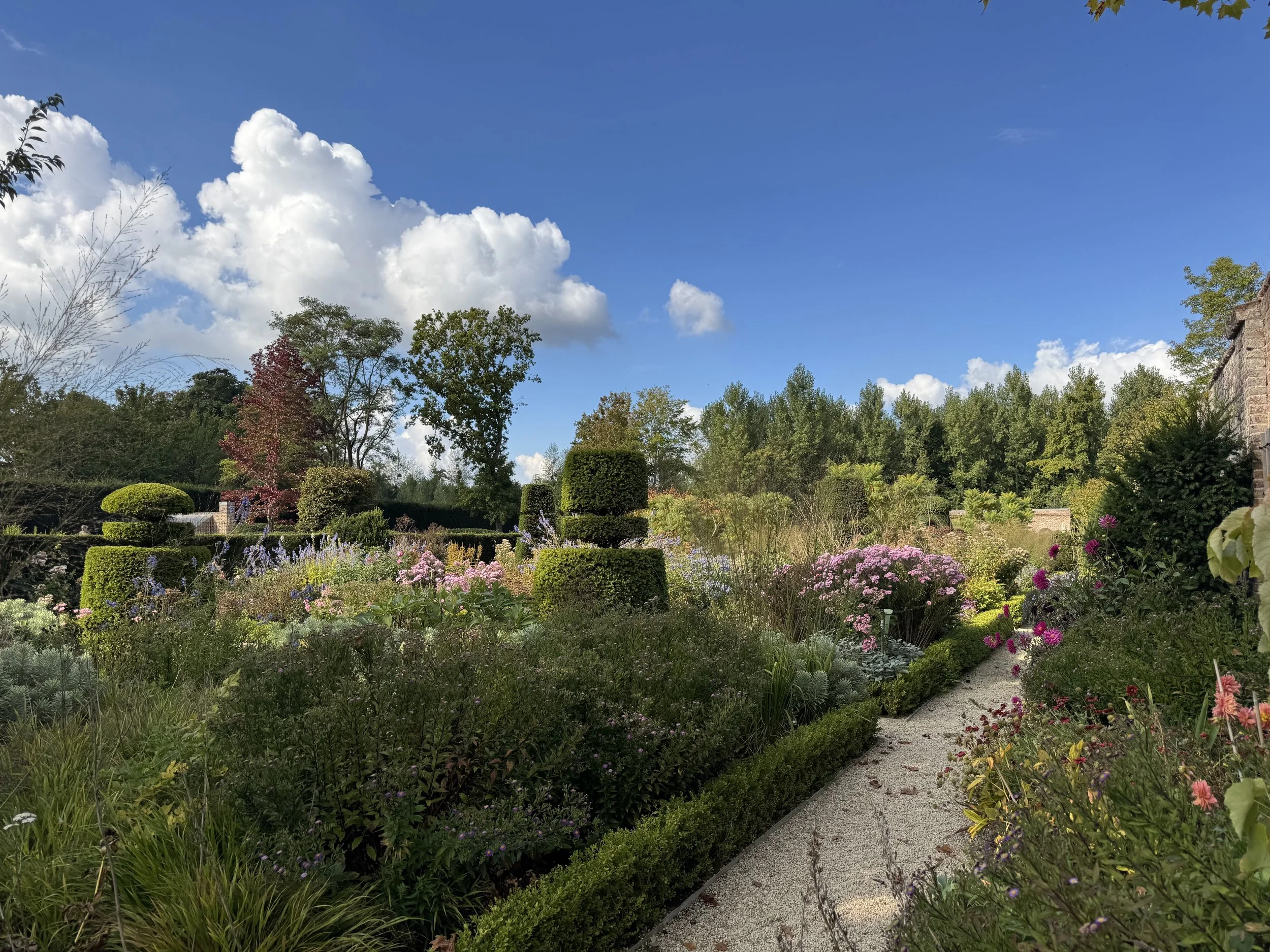 A colorful garden with flowering plants, trimmed bushes, and trees under a blue sky with white clouds.