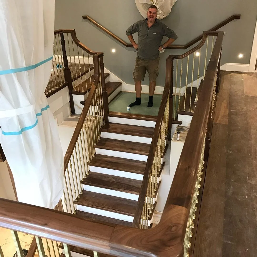 A man standing at the top of a staircase with a new wooden railing, smiling with his hands on his hips.