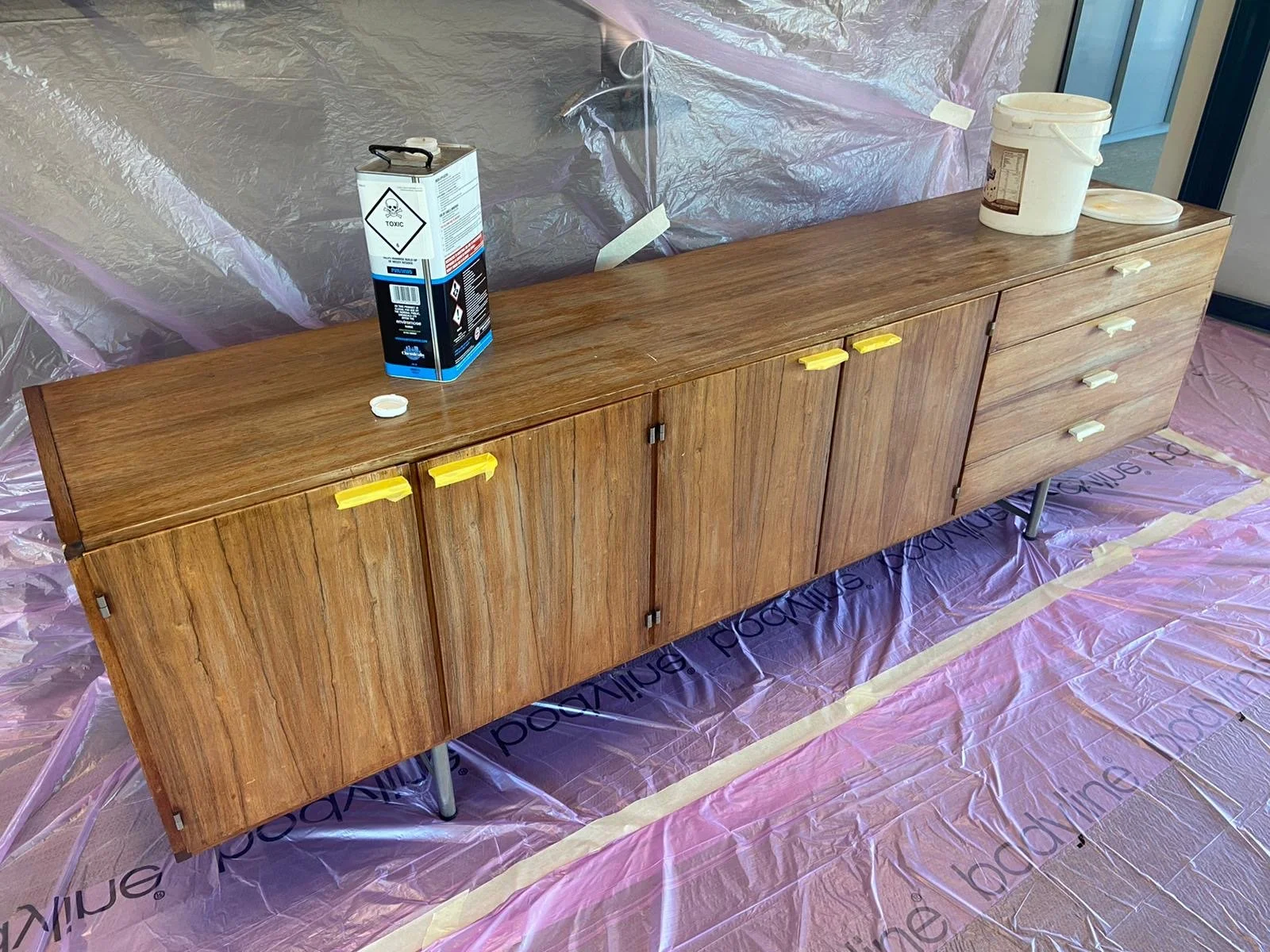 A wooden sideboard with yellow and white handles, placed on pink protective flooring with the brand name 'What's Painting'. On top of the sideboard, there is a can of toxic chemical, a white plastic container, and a small white lid.
