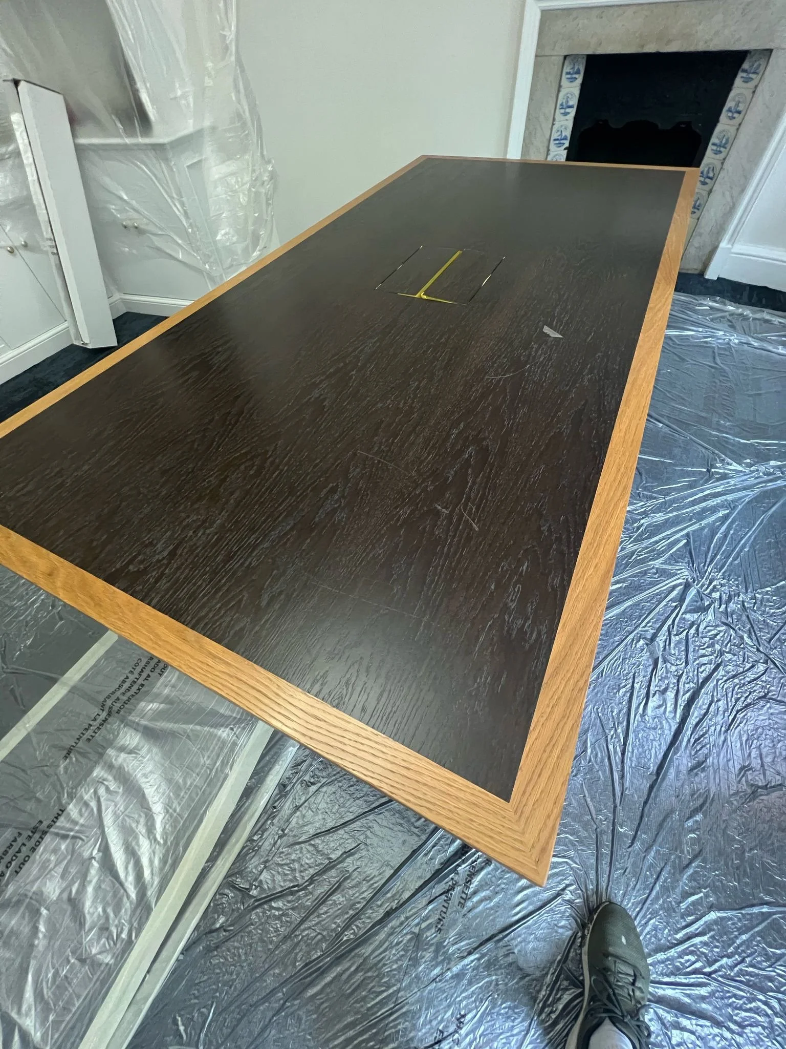A large wooden conference table with a dark top and lighter wood border, placed on protective plastic covering on the floor, with a fireplace in the background.