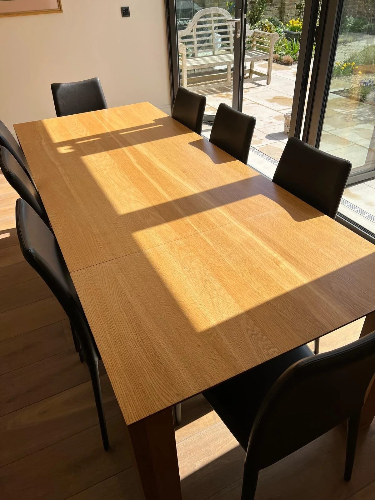 Wooden dining table with five black chairs, sunlight streaming in through large glass sliding doors showing an outdoor patio with garden furniture and flowers.