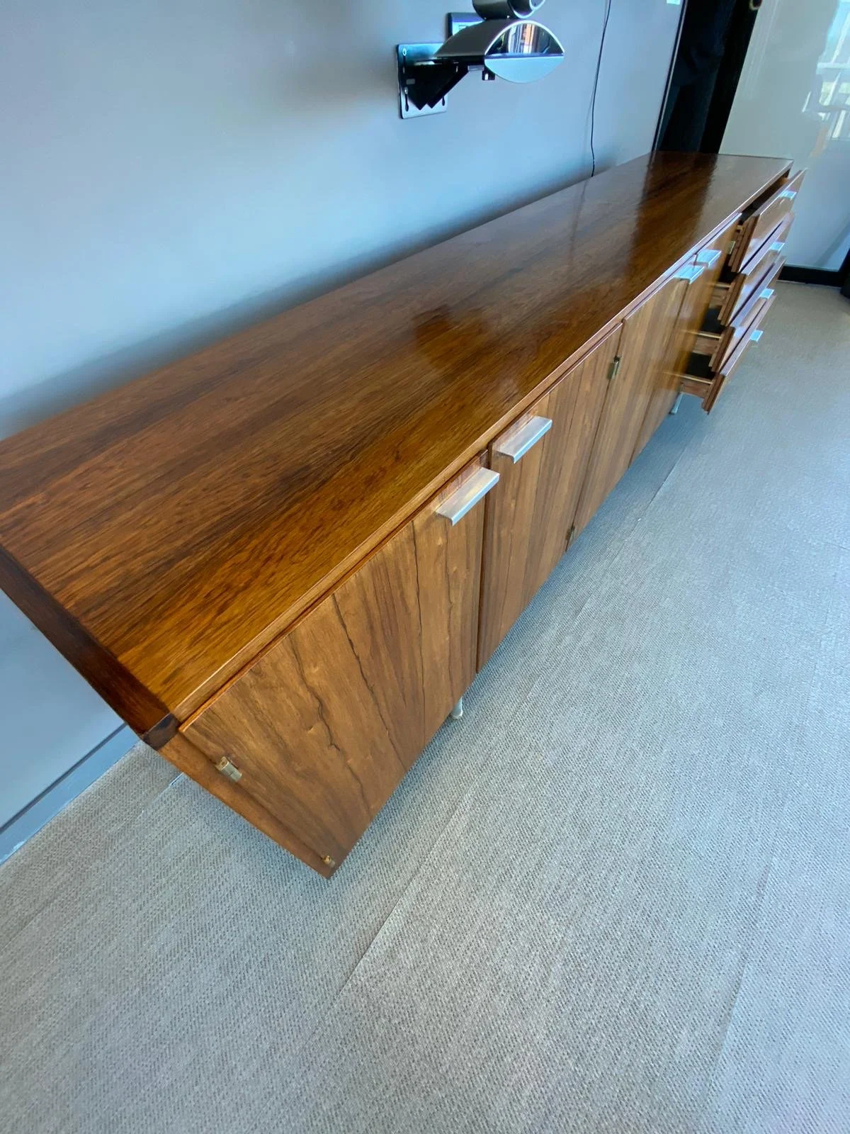 A wooden sideboard with multiple drawers and cabinets, placed against a light gray wall. A wall-mounted lamp is visible above the sideboard, and the floor is carpeted in beige.