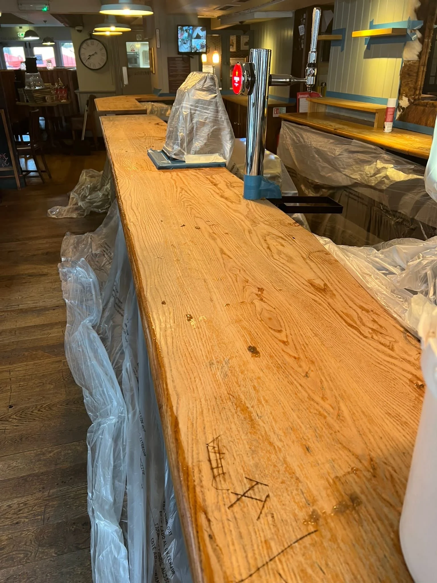 Wooden bar counter with plastic covering and some water droplets, in a restaurant or pub setting with tables, chairs, and wall decor in the background.