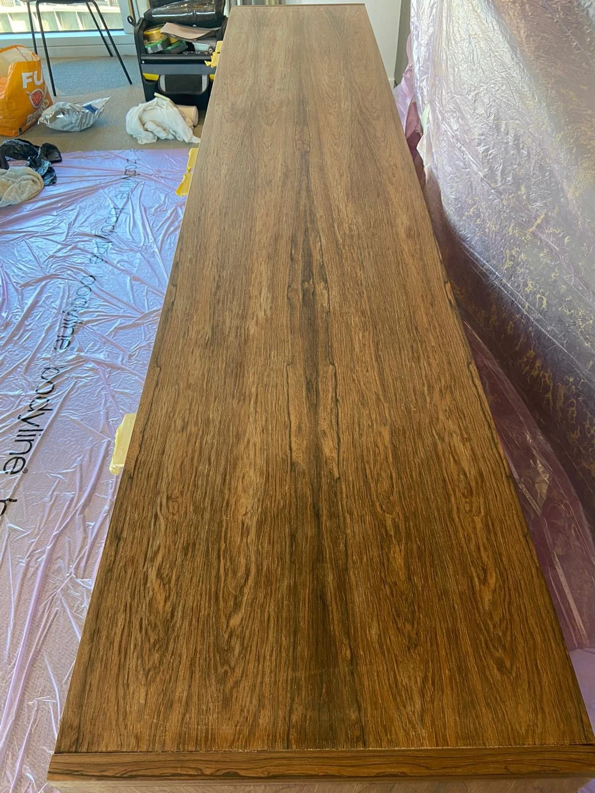 A long, wooden tabletop in a workshop or room with various tools, materials, and furniture around it.